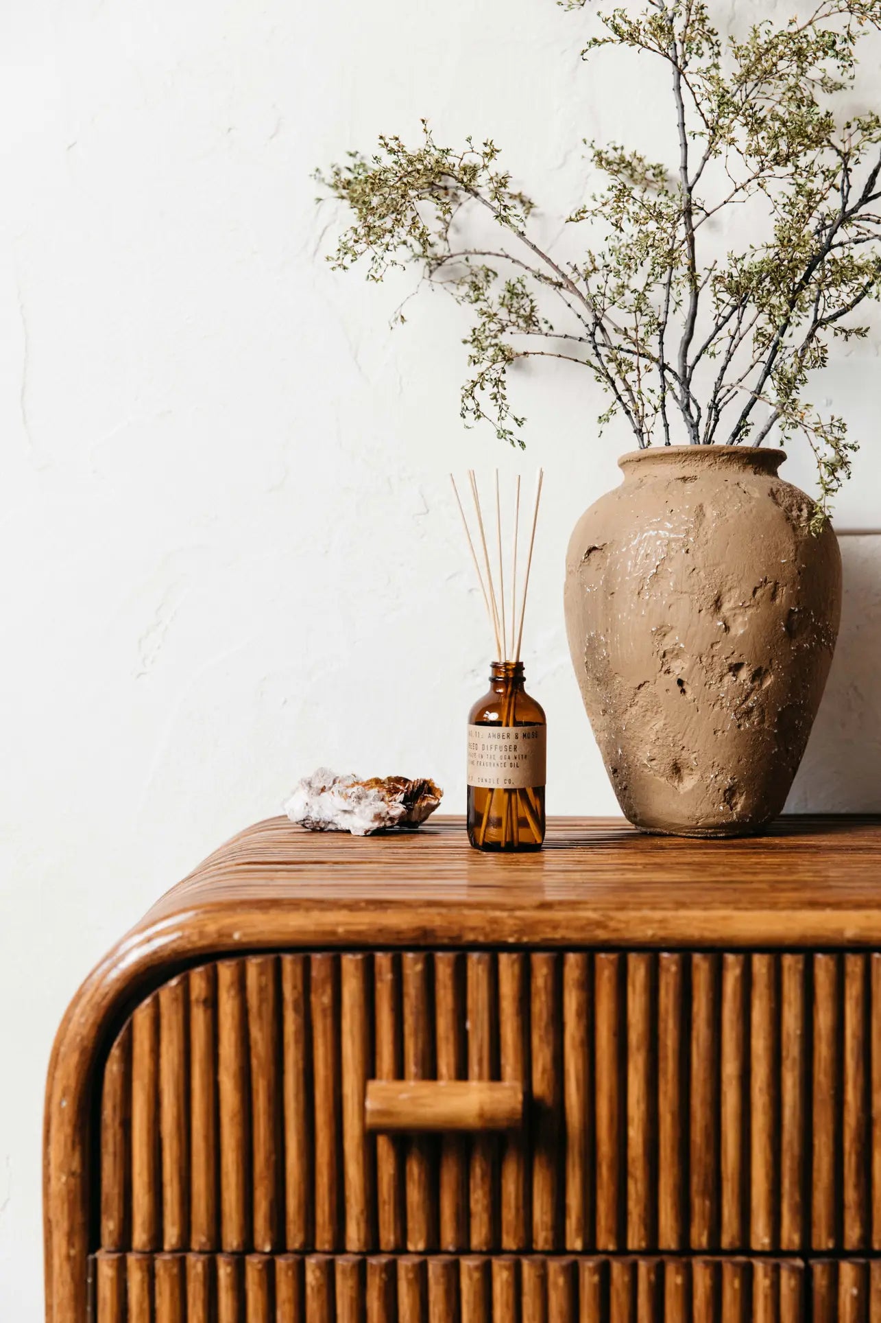 A brown ceramic vase with green foliage, a P.F. Candle Co. 3.5oz Reed Diffuser in Amber & Moss, and a crystal are set on a wooden dresser with a curved slatted front against a white textured wall.