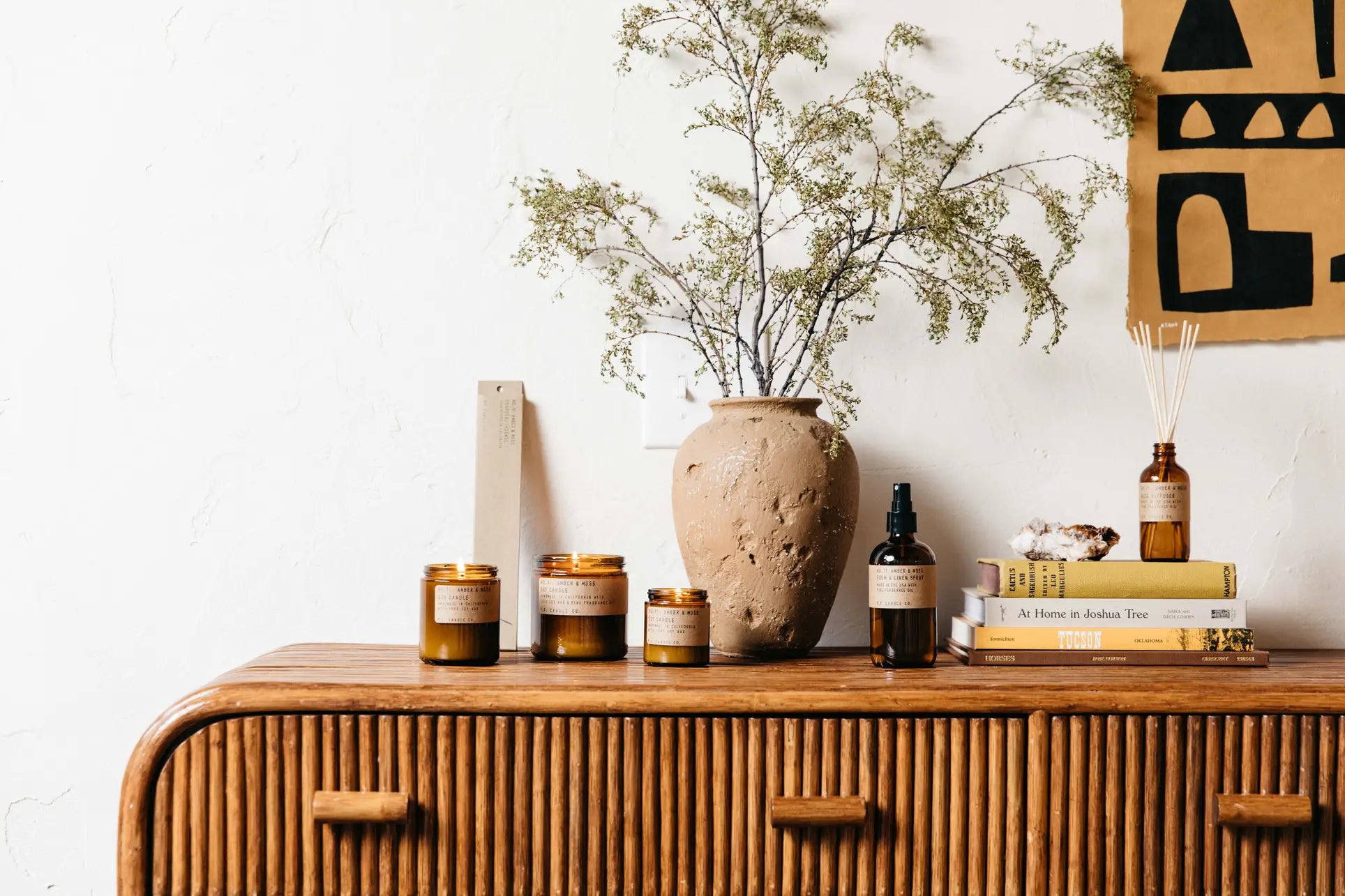 A wooden sideboard displays amber glass candles, a rustic vase with green branches, P.F. Candle Co. 3.5oz Reed Diffuser in Amber & Moss, books, a spray bottle, and sits against a white wall with abstract art.