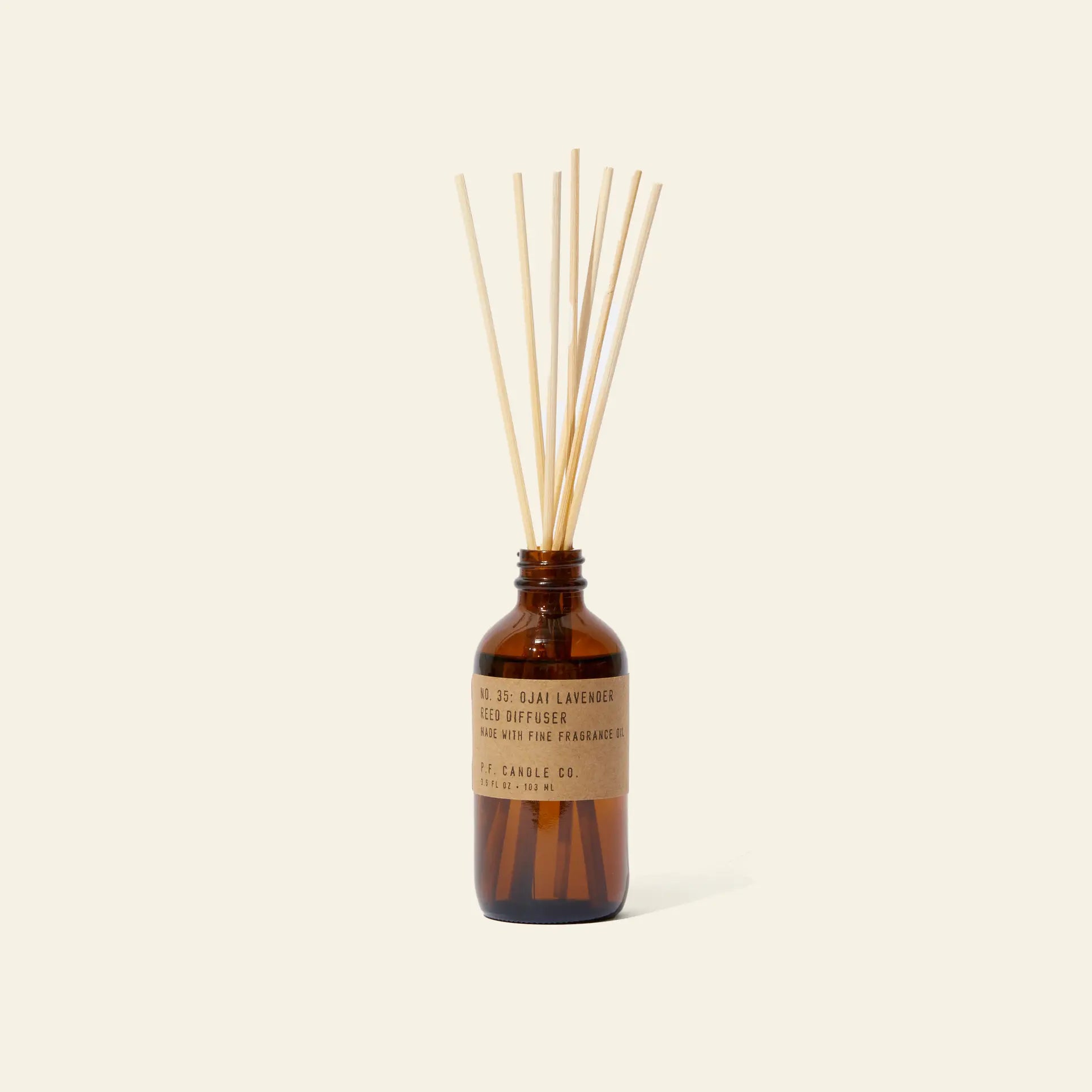 The P.F. Candle Co. 3.5oz Reed Diffuser in Ojai Lavender features several natural reeds in a brown glass bottle with a tan label, releasing soothing fragrance oils into the air against a plain light background.