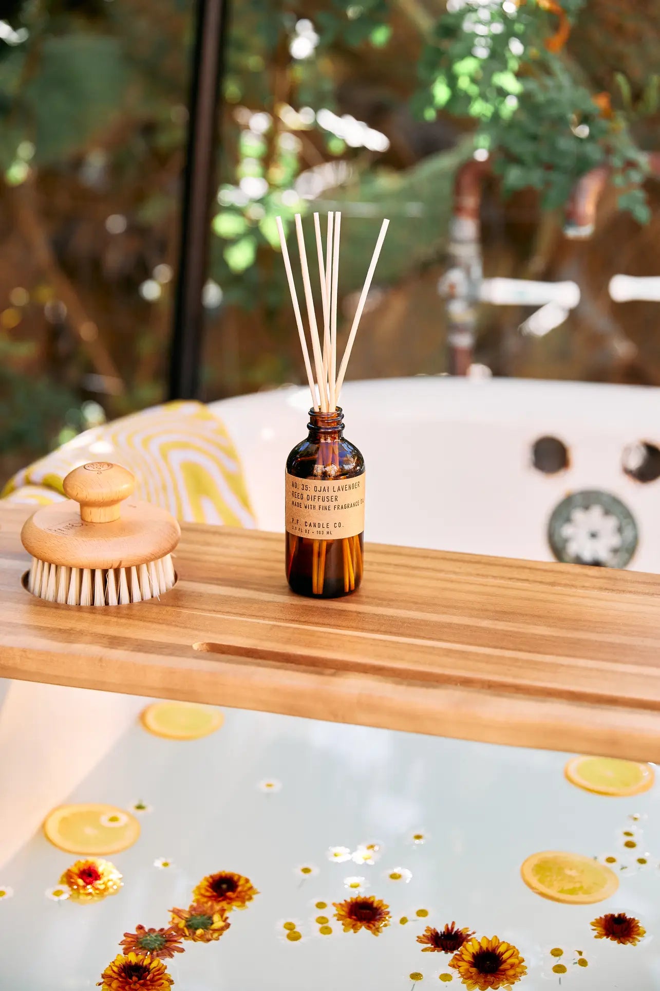 A wooden bath tray holds a round dry brush and the P.F. Candle Co. 3.5oz Reed Diffuser in Ojai Lavender, filling the air with fragrance above a tub with floating citrus slices and orange flowers. Greenery can be seen outside the window.