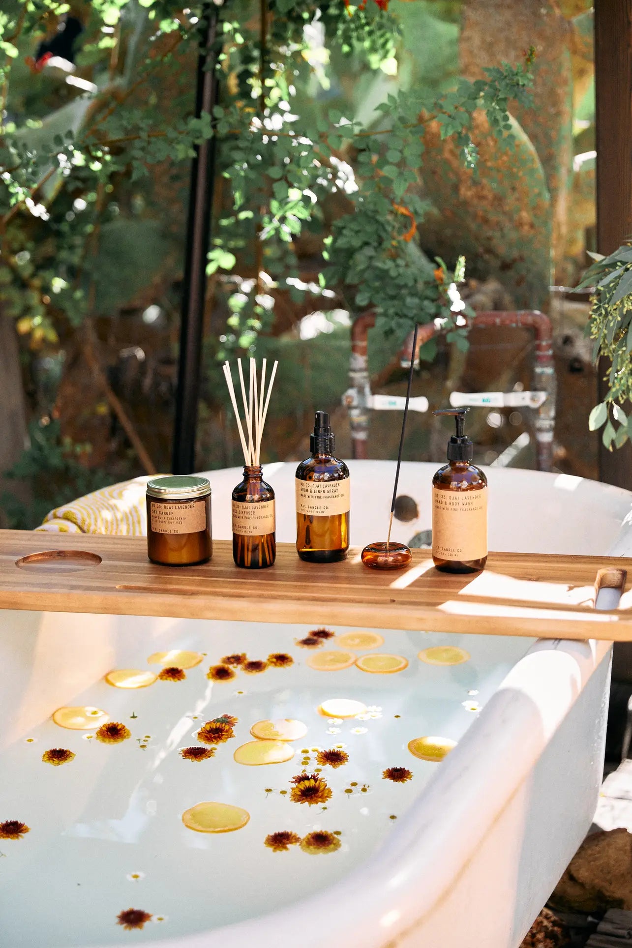 A white bathtub filled with water, lemon slices, and petals sits outdoors among greenery. A wooden tray features the P.F. Candle Co. 3.5oz Reed Diffuser - Ojai Lavender, candles, amber bottles, and fragrance oils for an elevated aromatic experience.