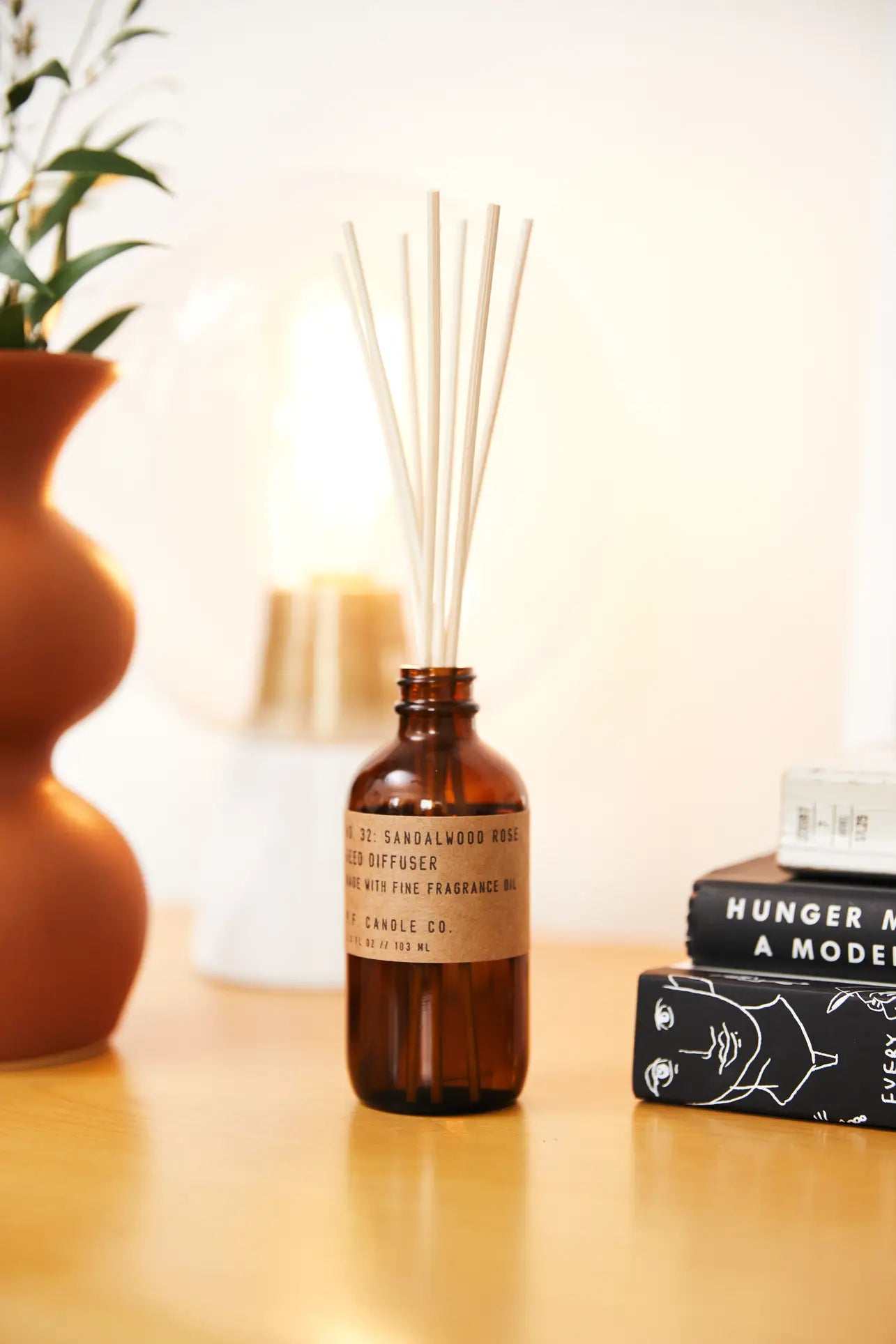 A P.F. Candle Co. 3.5oz Reed Diffuser in Sandalwood Rose sits on a wooden surface next to a brown vase, a lamp, and a stack of books including one titled Hunger Makes Me a Modern Girl.