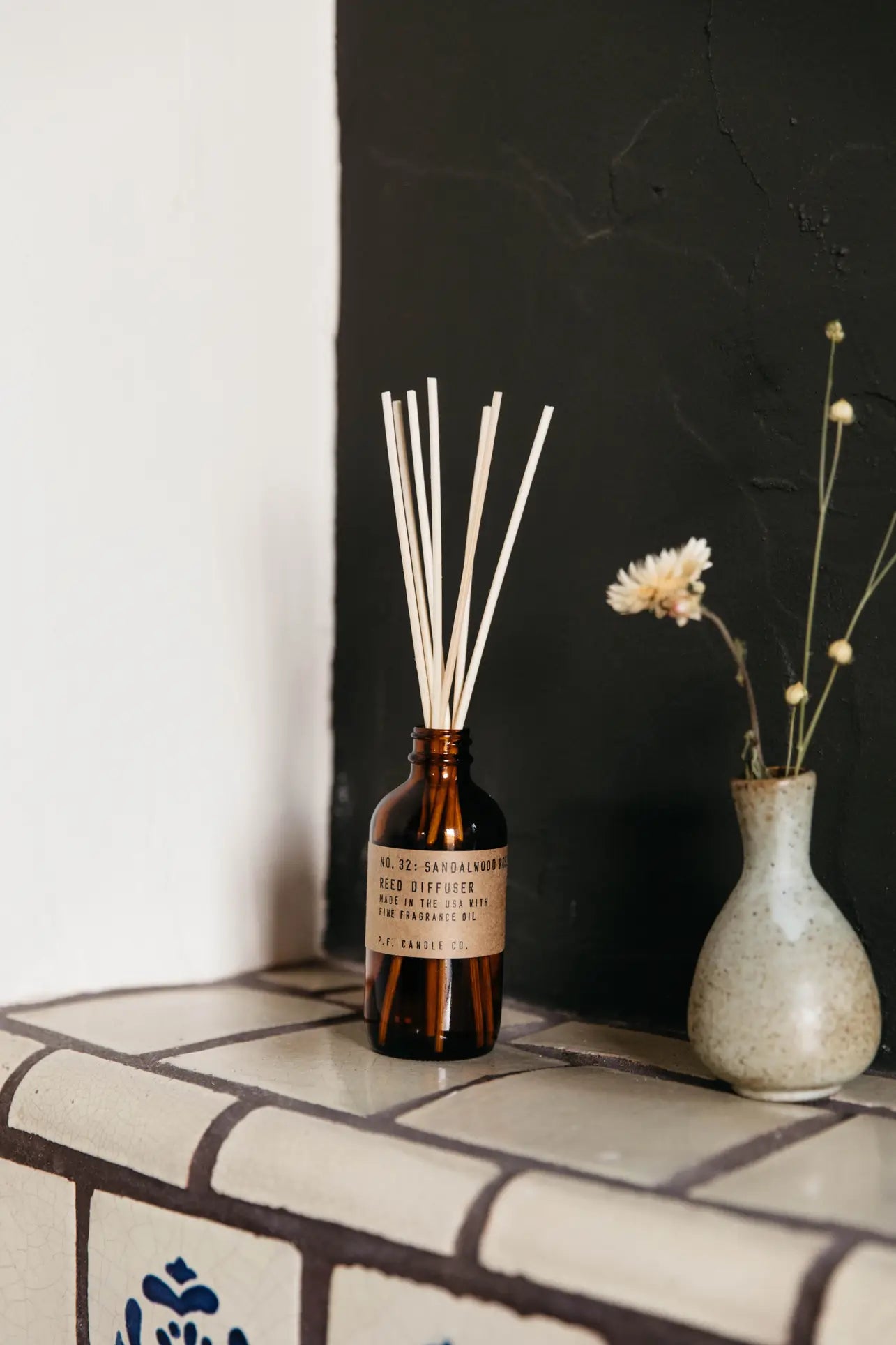 The P.F. Candle Co. 3.5oz Reed Diffuser in Sandalwood Rose adds fragrance on a tiled surface beside a small vase of dried flowers, set before a black and white wall.