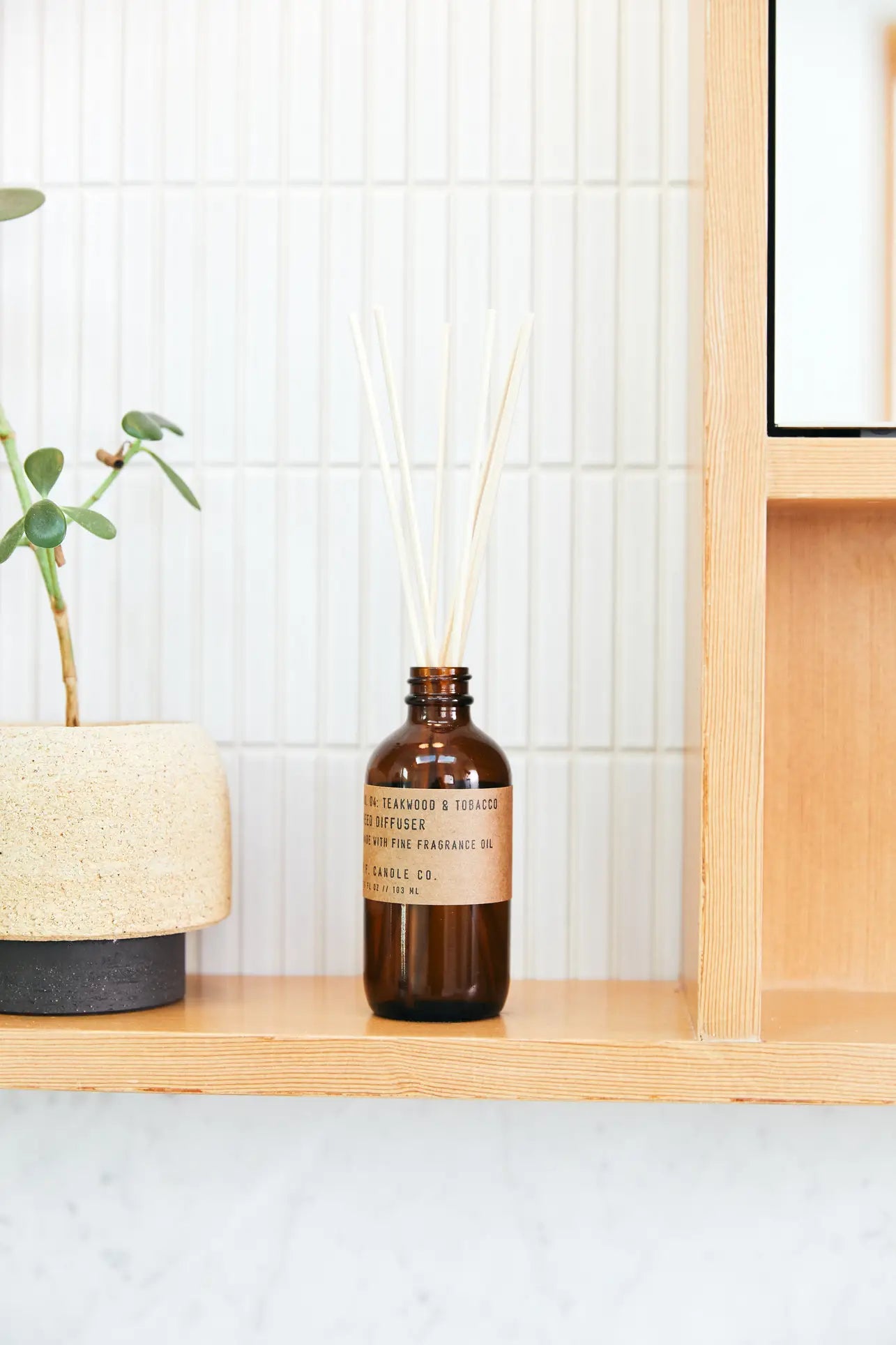 The P.F. Candle Co. 3.5oz Reed Diffuser in Teakwood & Tobacco, with several sticks, sits on a wooden shelf near a beige potted plant against a white tiled wall, filling the space with high-quality fragrance oils.