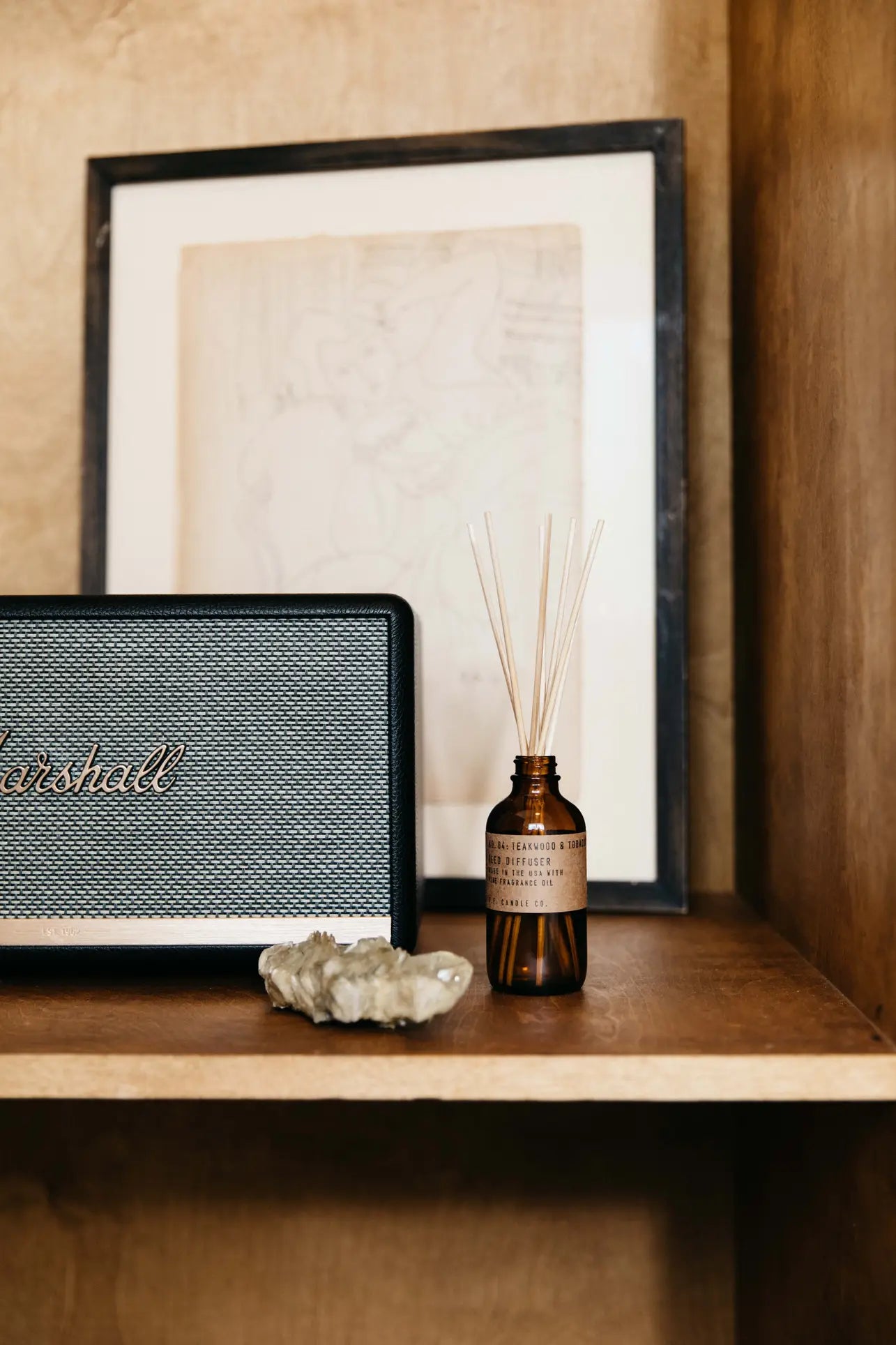A close-up of a shelf with a Marshall speaker, crystals, and a P.F. Candle Co. 3.5oz Reed Diffuser in Teakwood & Tobacco, next to a framed sketch in a warm-toned minimalist setting.