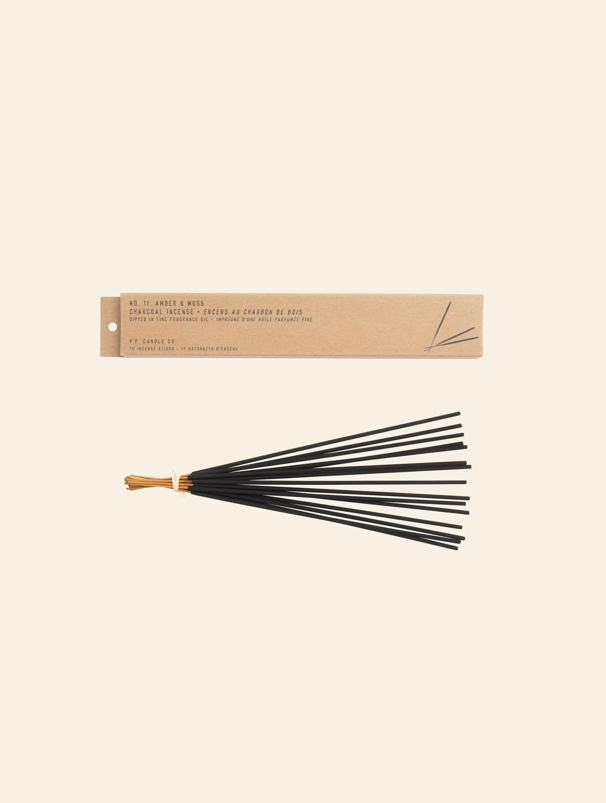 A bundle of black incense sticks tied with a tan string rests below a rectangular P.F. Candle Co. Classic Incense package labeled No. 11 Amber & Moss, evoking subtle notes of teakwood on a simple light background.