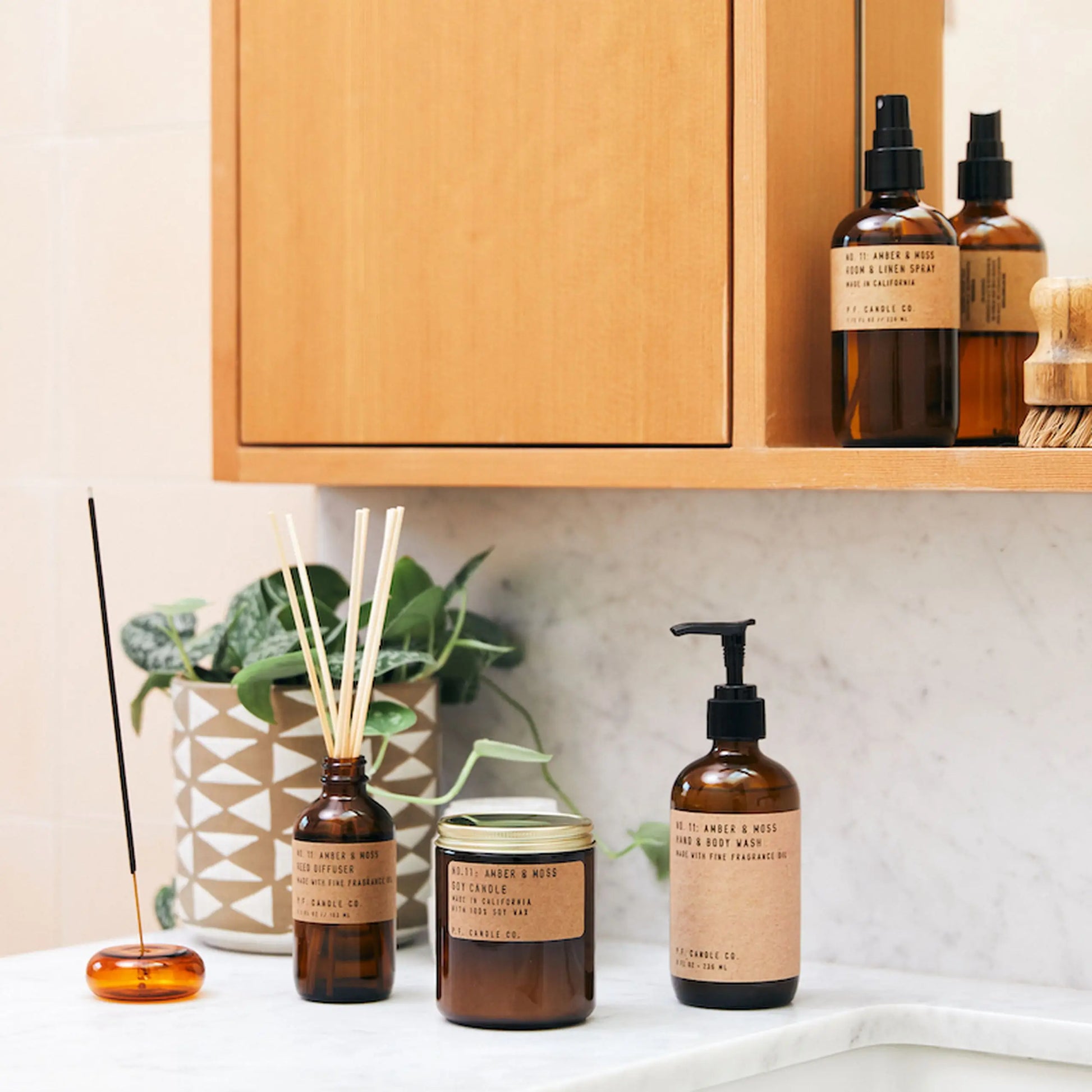 P.F. Candle Co. No. 11 Amber & Moss Soy Candle, amber glass bottles with brown labels, a reed diffuser, and a pump soap bottle are neatly arranged on the bathroom counter beside a plant and fragrant incense smoke.