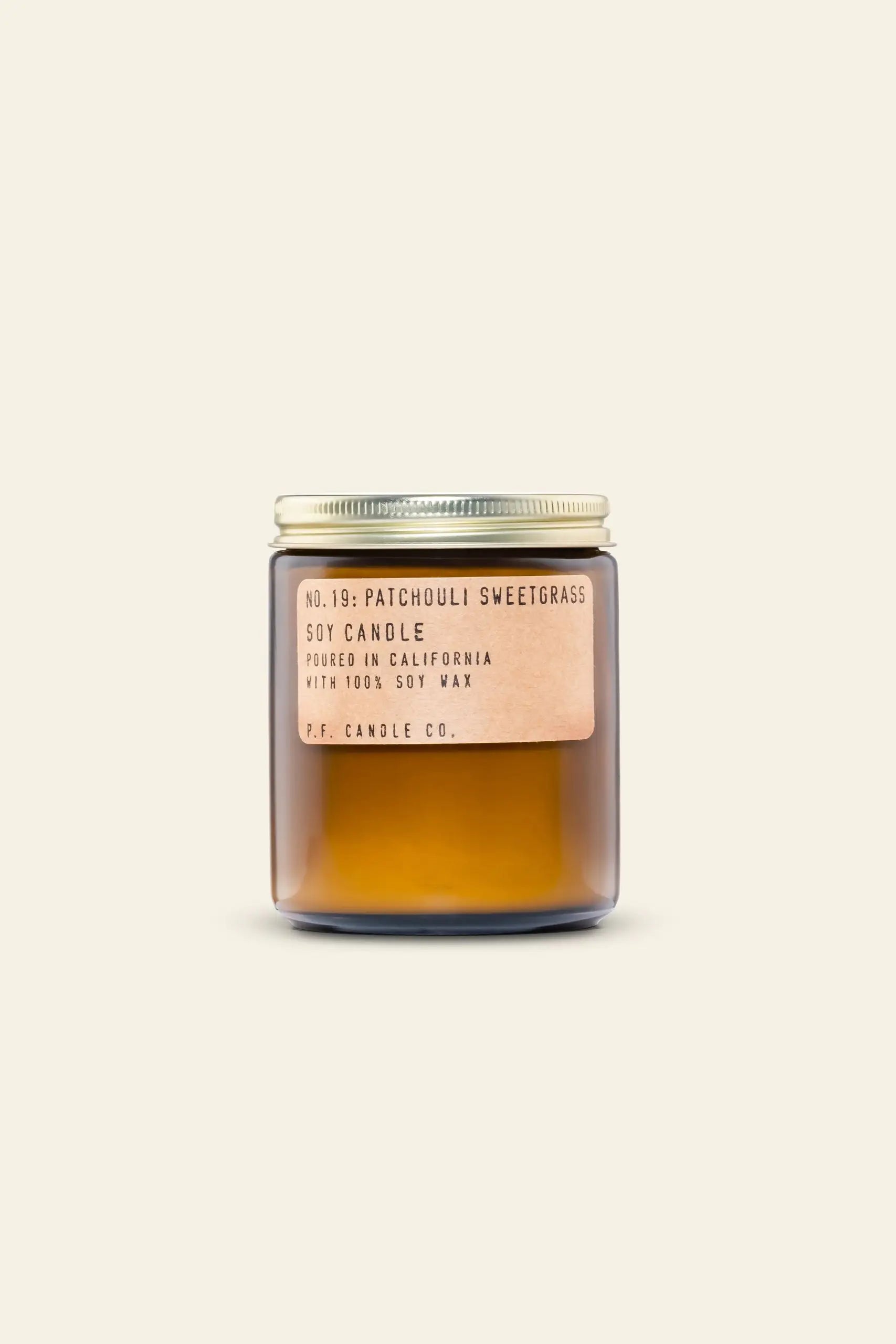 The P.F. Candle Co. No. 19 Patchouli Sweetgrass 7.2 oz Soy Candle features a brown glass jar, gold metal lid, and tan label—crafted with clean-burning soy wax for a calming atmosphere. Shown on a plain beige background.