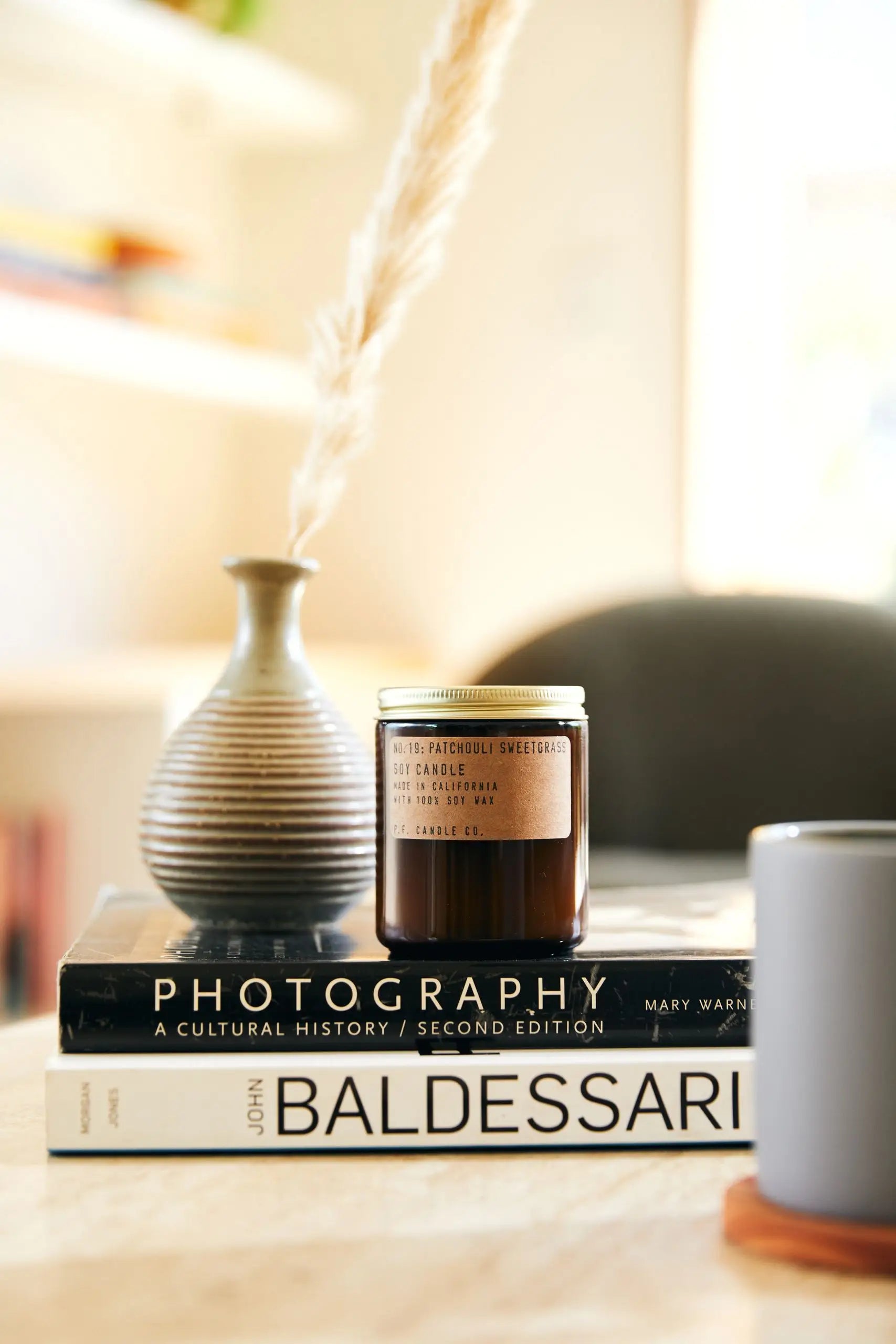 A P.F. Candle Co. No. 19 Patchouli Sweetgrass 7.2 oz Soy Candle rests on two books beside a striped ceramic vase with pampas grass; a gray mug is partially visible, creating an inviting scene for relaxation and reflection.
