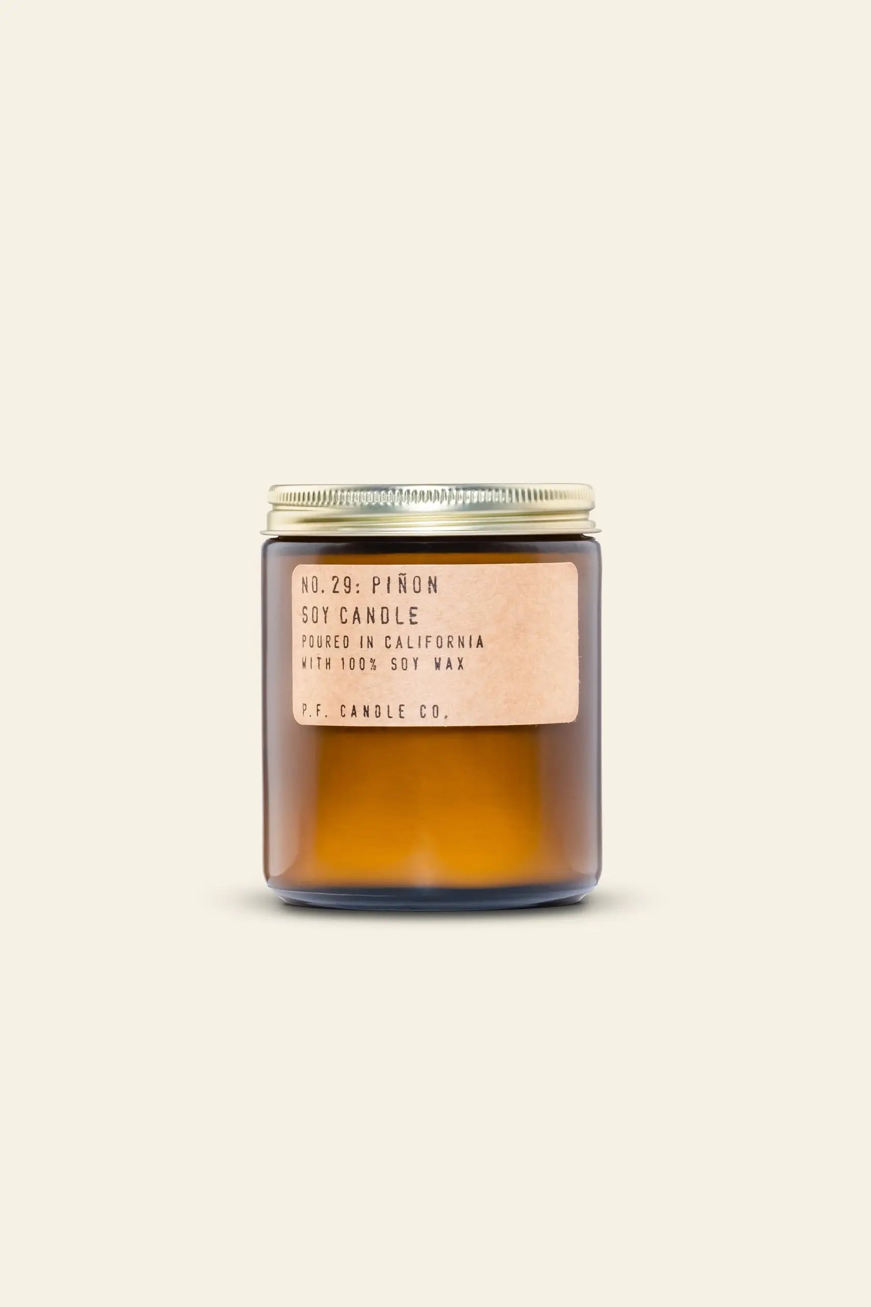 A brown glass jar candle with a gold lid and beige label reading No. 29: Piñon, poured in California with 100% soy wax by P.F. Candle Co., featuring warm cedarwood notes. Background is plain light cream. Product: P.F. Candle Co. No. 29 Piñon 7.2 oz Soy Candle.