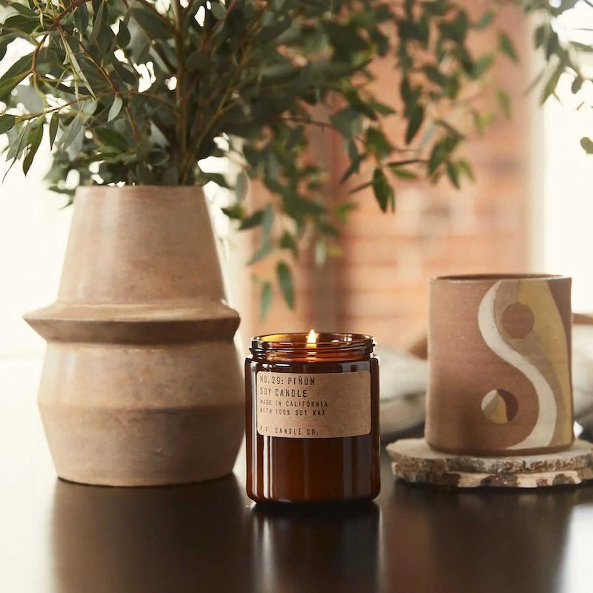 A P.F. Candle Co. No. 29 Piñon 7.2 oz Soy Candle in amber glass glows on a dark table next to a tan ceramic vase with greenery and a decorative container, its cedarwood notes bringing warmth to the cozy setting.