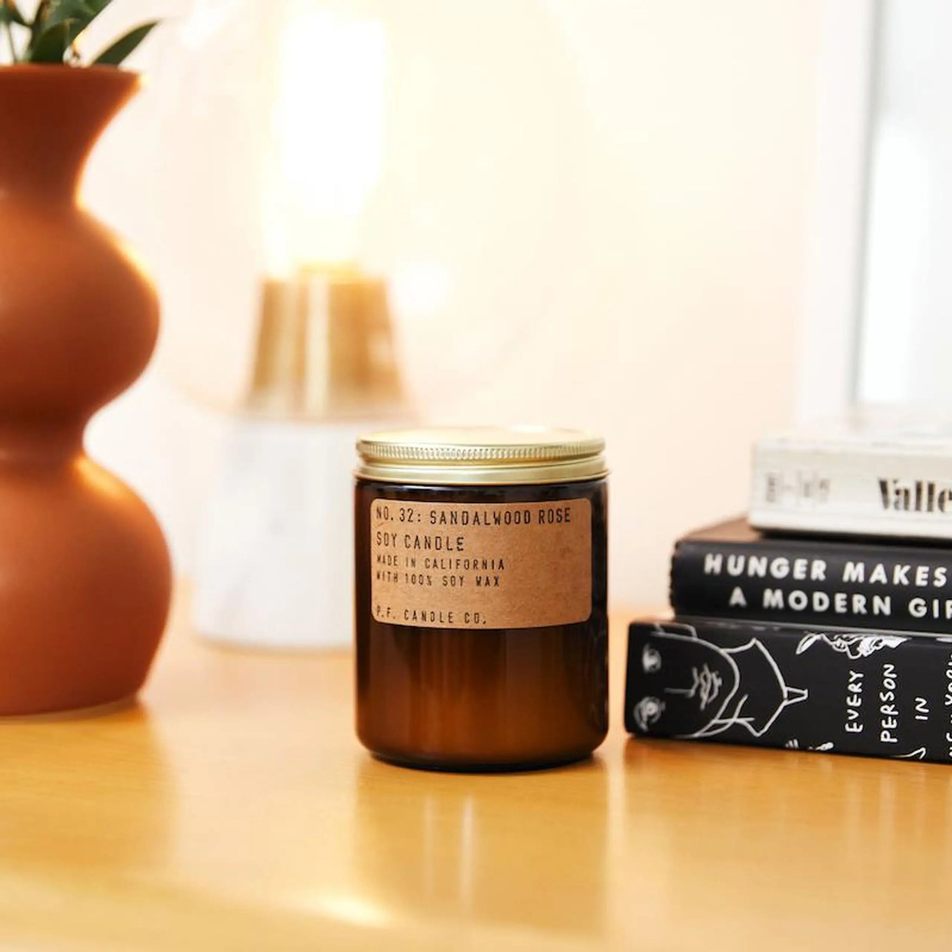 The P.F. Candle Co. No. 32 Sandalwood Rose 7.2 oz Soy Candle sits on a wooden surface next to stacked books, a ceramic vase with greenery, and a softly blurred lamp in the background.