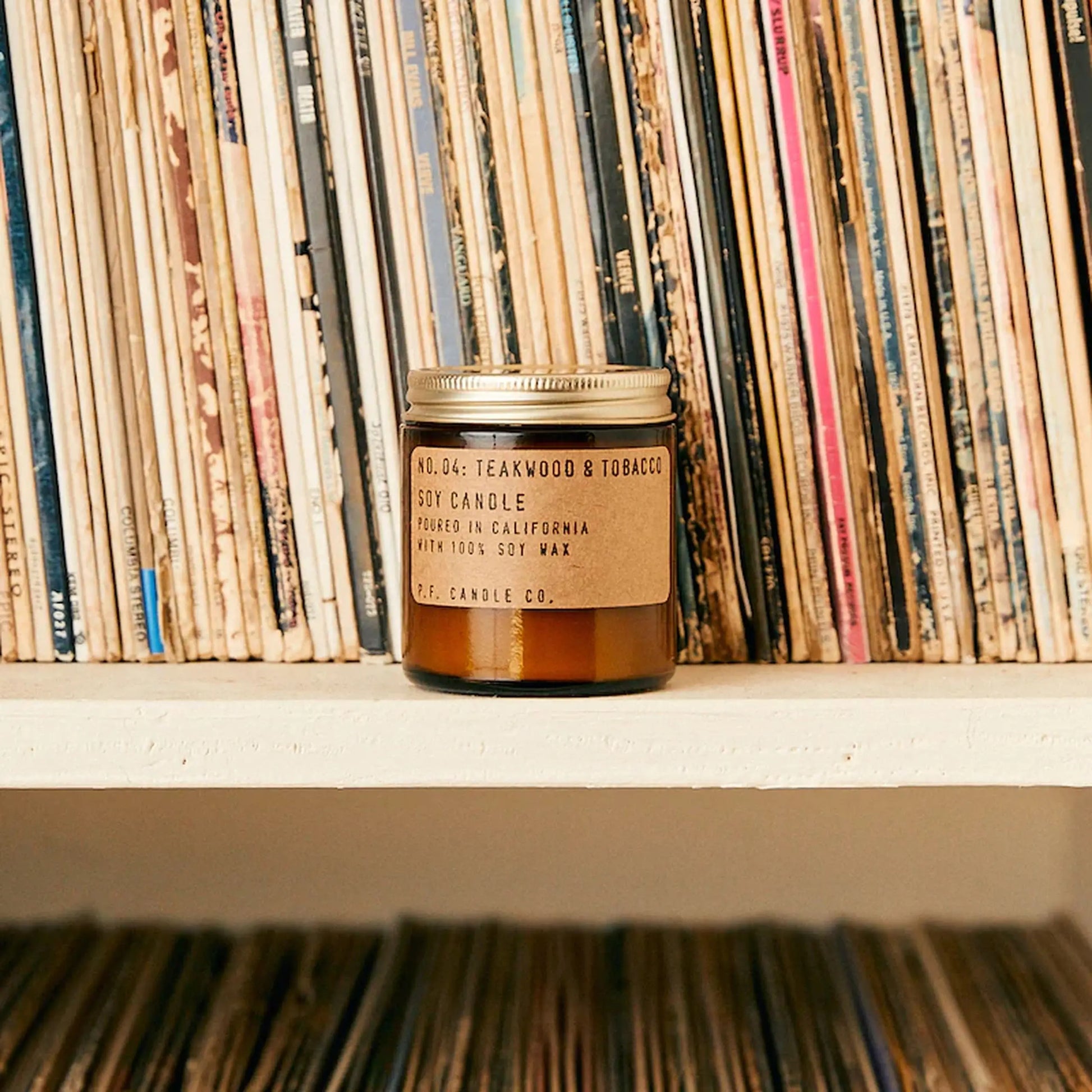 A P.F. Candle Co. No. 4 Teakwood & Tobacco 3.5 oz Mini Candle rests on a white shelf in front of vinyl records with colorful spines.