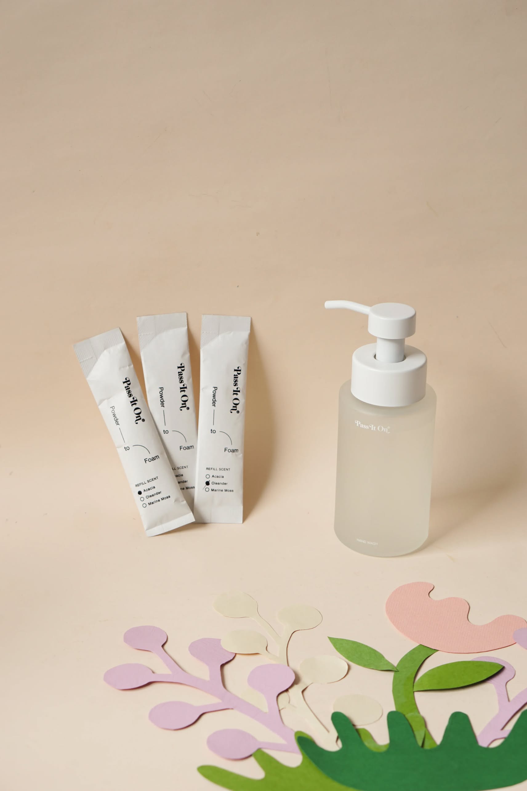 A minimalist photo displays three Pass It On 0.02 Refillable Hand Wash Set sachets in Marine Moss and a frosted pump bottle, with pastel paper leaves arranged in the foreground on a neutral background.