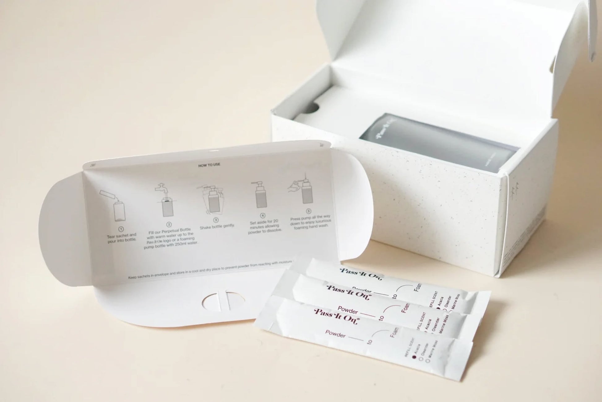 A white box with an open lid reveals the Pass It On 0.02 Refillable Hand Wash Set, Pack of 3-Oleander. In front are two single-use packets and an illustrated instruction card from Pass It On, highlighting botanically-derived, sustainable hand wash.