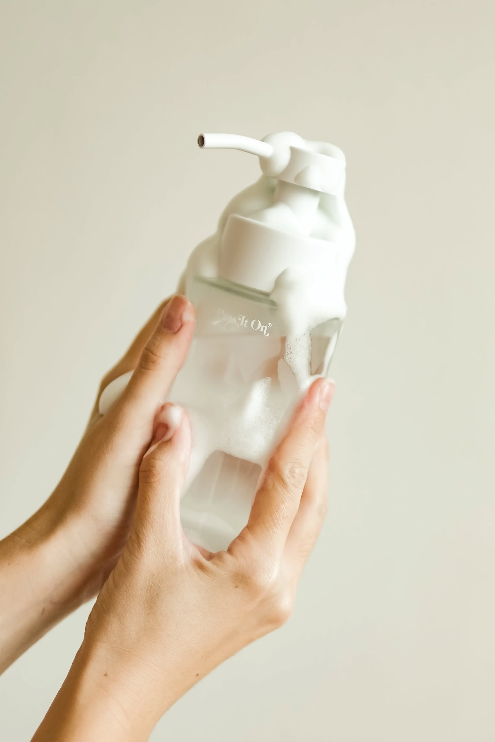 Two hands hold Pass It On 0.02 Refillable Hand Wash Set, Pack of 3-Oleander, a sustainable, botanically-derived hand wash by Pass It On. The transparent pump bottle is partially obscured by white foam against a plain background.