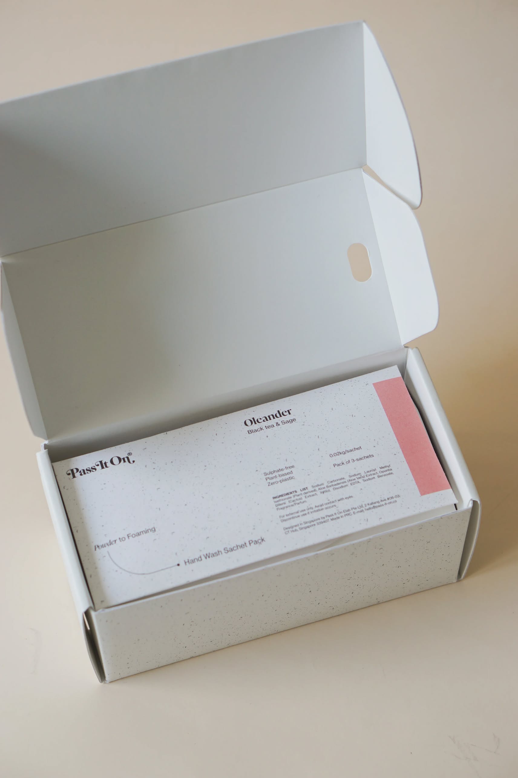 A white rectangular box from Pass It On sits open on a light surface, revealing neatly arranged cards. The top card reads Pass It On 0.02 Refillable Hand Wash Set, Pack of 3-Oleander, a sustainable hand wash sachet pack.