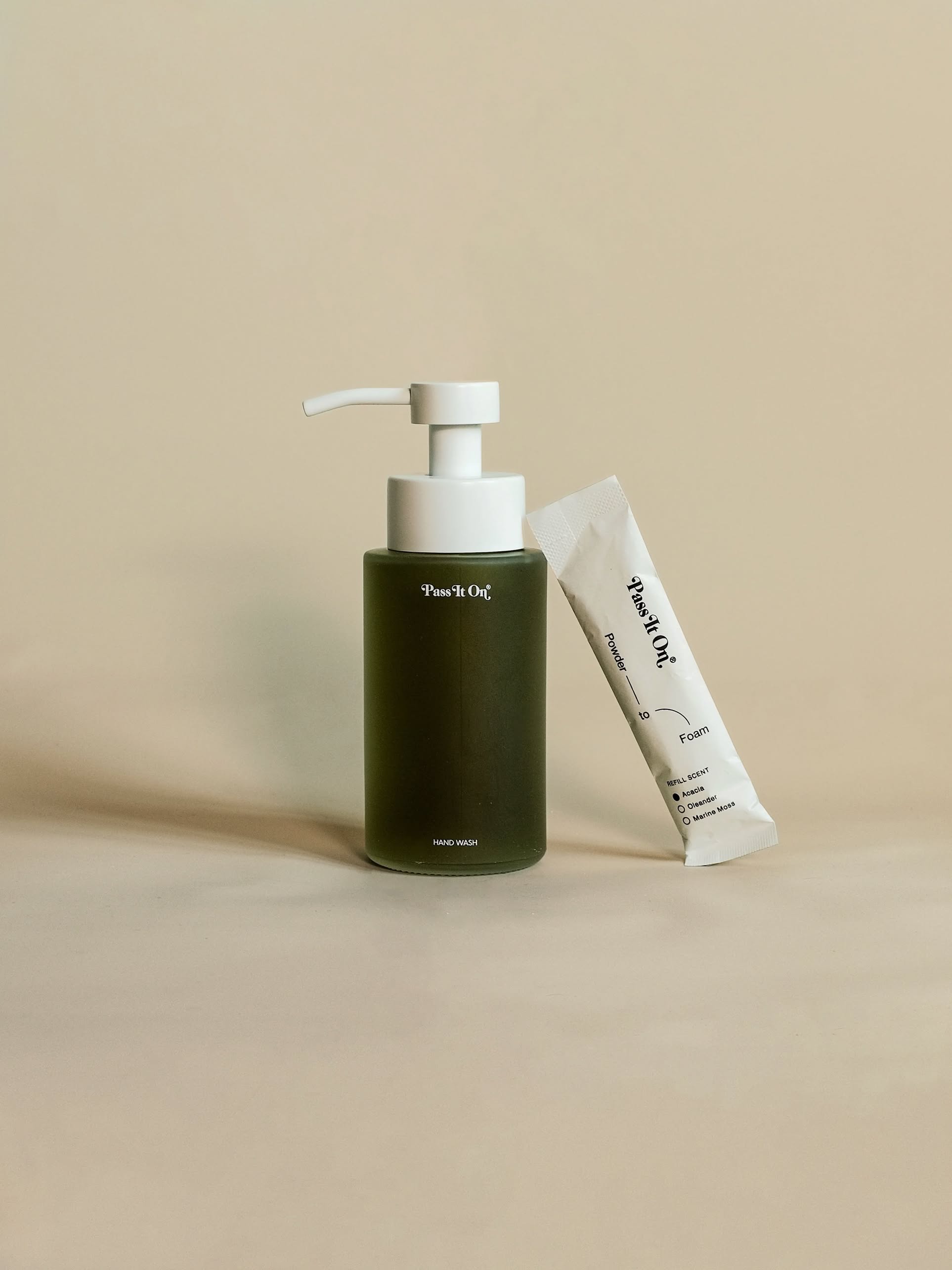 A green pump bottle of Pass It On 0.02 Refillable Hand Wash Set, Single Pack - Acacia stands beside a white “Pour le Oui” sachet on a beige background, suggesting a refreshing cedarwood scent.