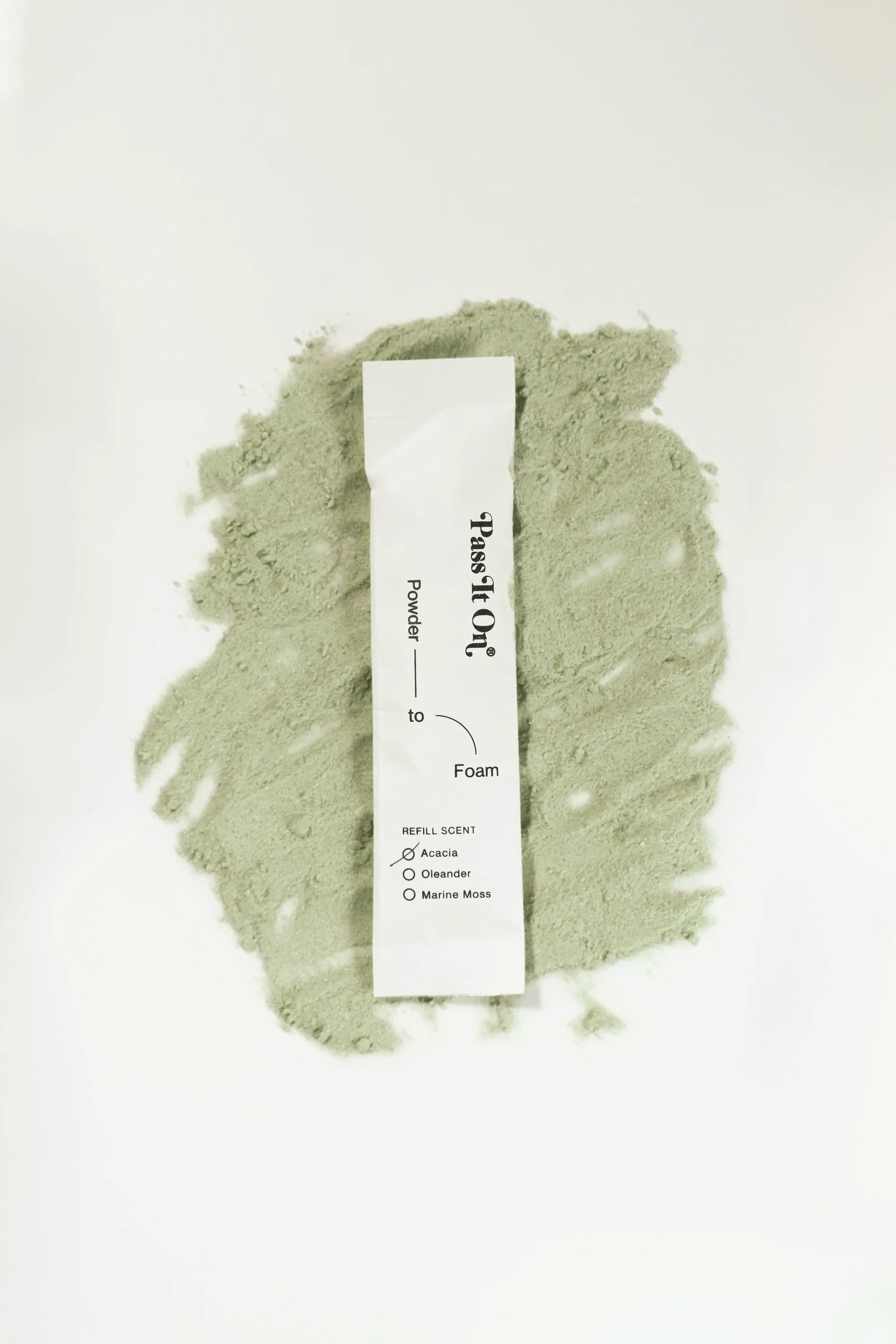 A white Pass It On 0.02 Refillable Hand Wash Set, Single Pack - Acacia, rests on light green powder, with scent options like Acacia, Osmanthus, Marine Moss, and cedarwood printed on the refillable packet.