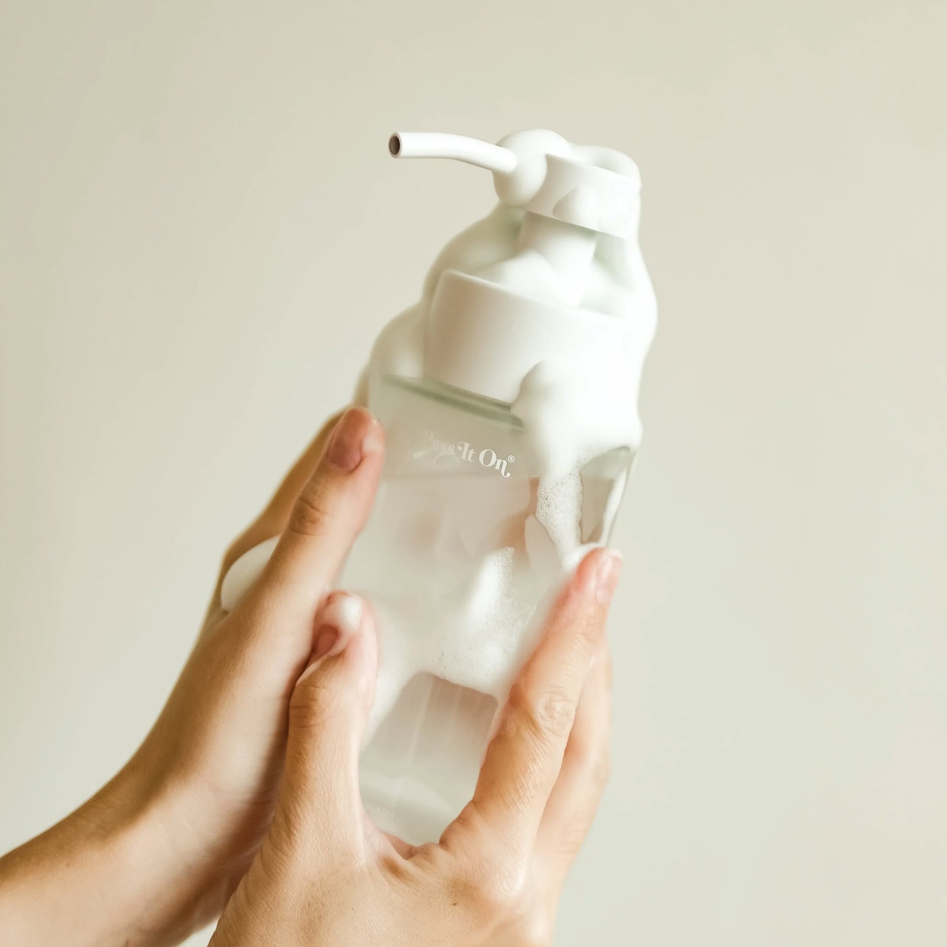 Hands holding the Pass It On 0.02 Refillable Hand Wash Set, Single Pack - Acacia pump bottle, covered in white foamy soap, against a light beige background.
