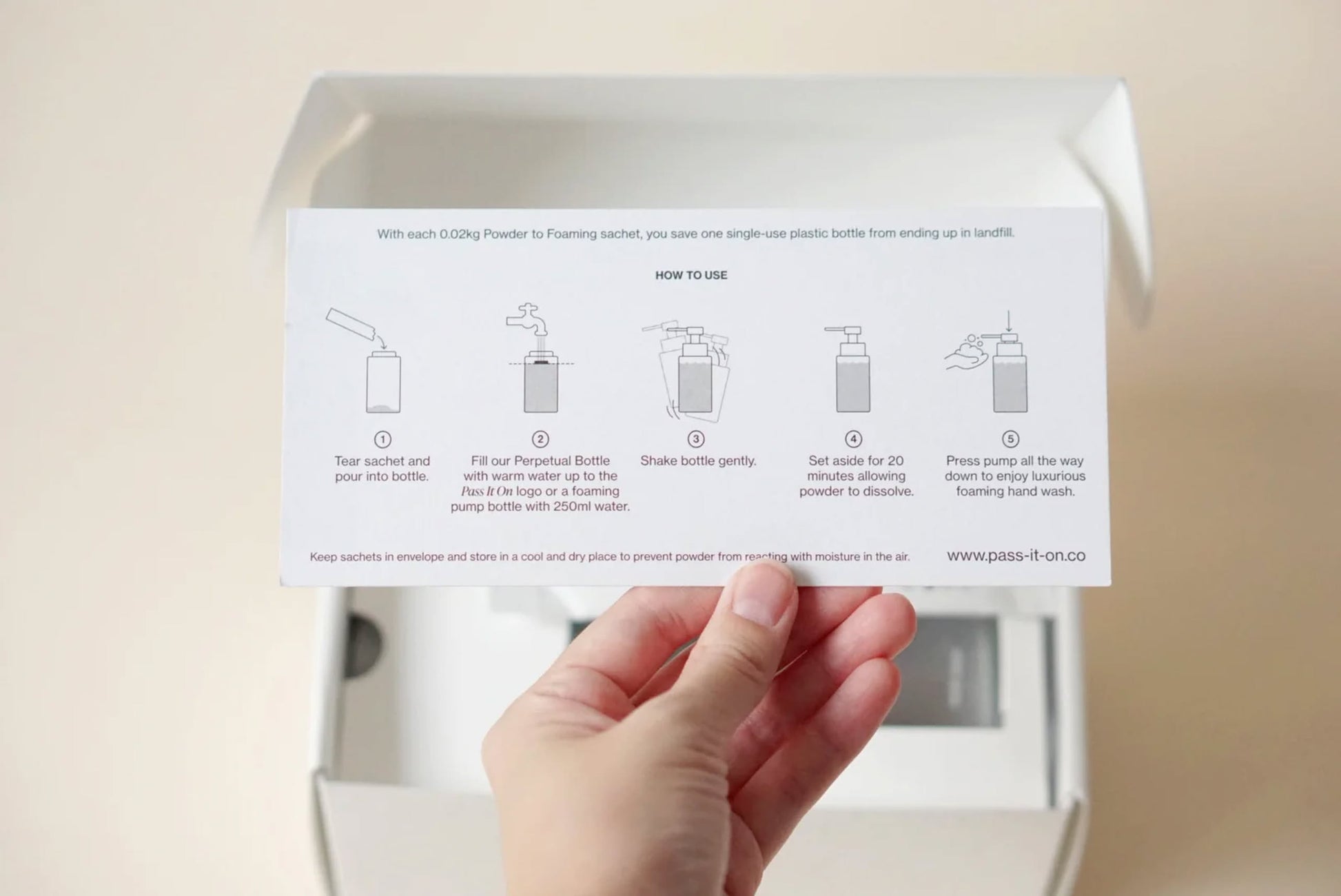 A hand holds an instruction card showing how to mix Pass It On 0.02 Refillable Hand Wash Set, Single Pack - Acacia with water in a bottle. Illustrated steps explain how to tear, fill, shake, wait, and use as the product box sits in the background.