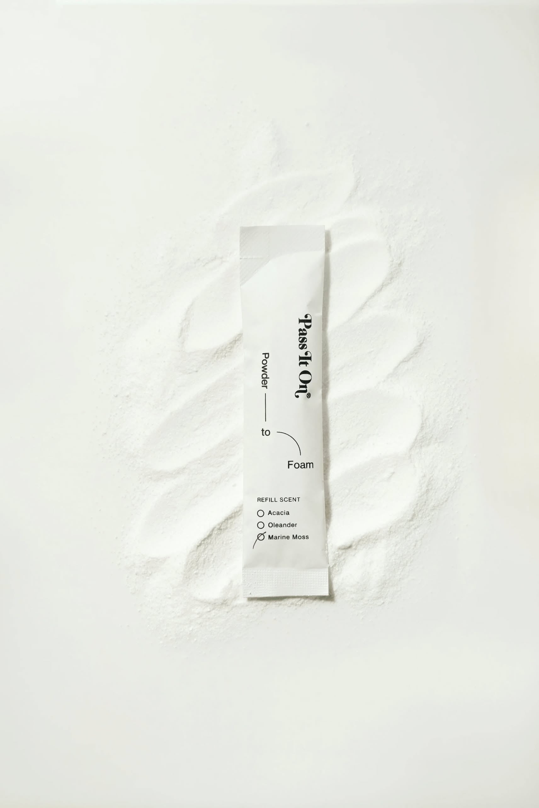A white sachet labeled Pass It On 0.02 Refillable Hand Wash Set, Single Pack - Marine Moss rests on white powder, highlighting its sustainable, refillable design by Pass It On against a clean, minimal background.