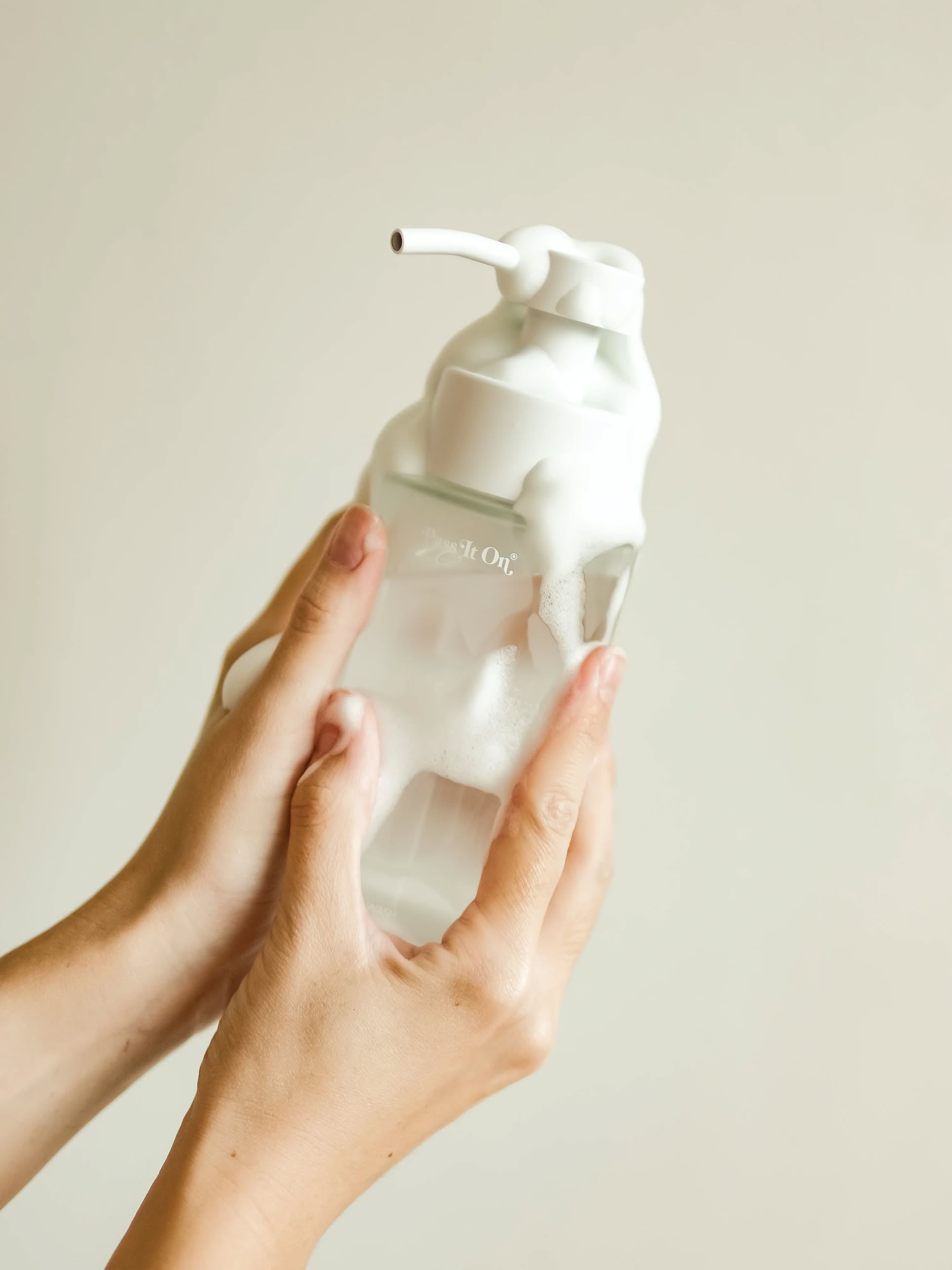 Two hands hold the Pass It On 0.02 Refillable Hand Wash Set, Single Pack - Marine Moss. White foam covers the clear dispenser, set against a plain light beige background highlighting this sustainable, gentle cleansing solution from Pass It On.