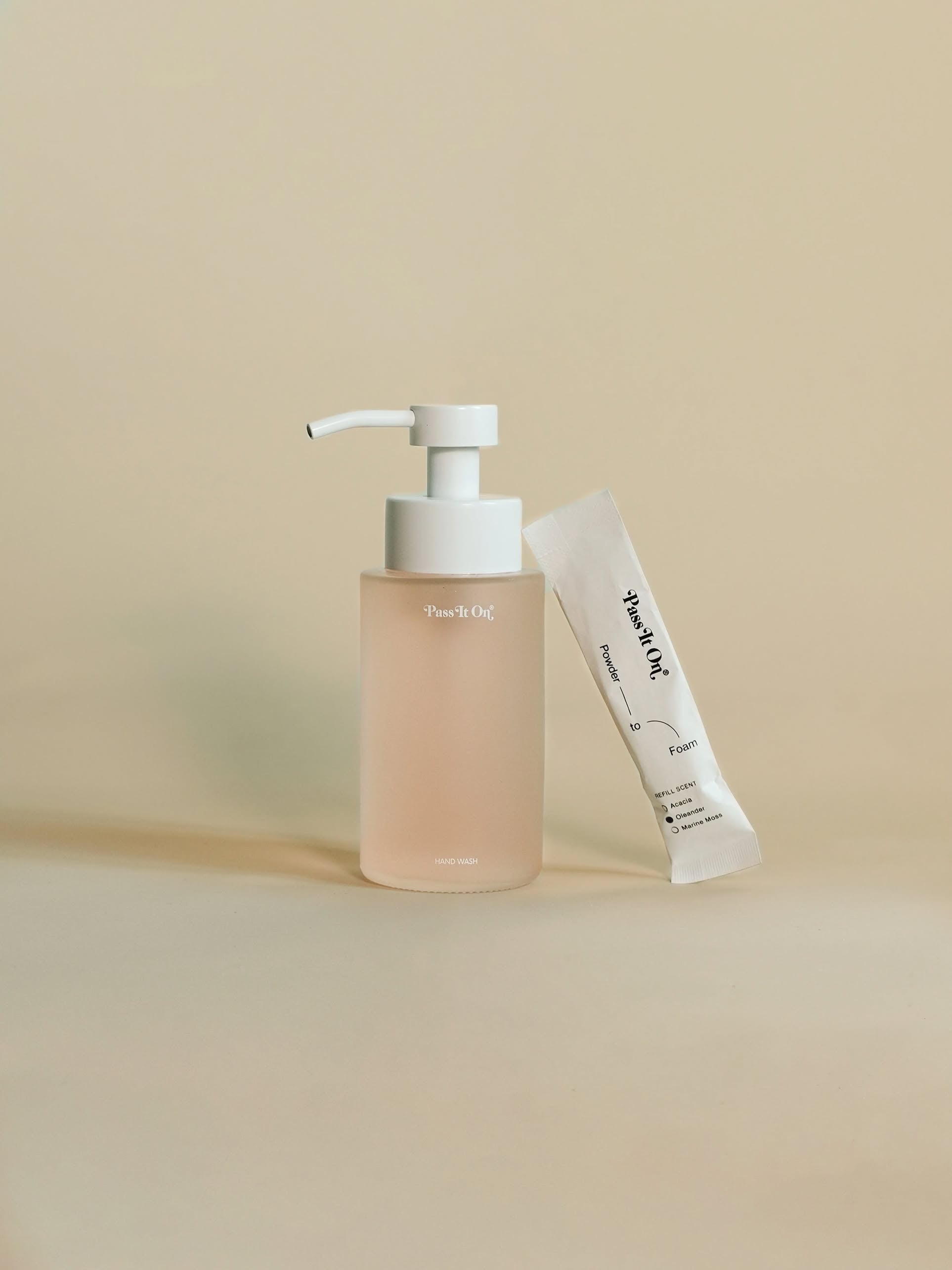 The Pass It On 0.02 Refillable Hand Wash Set, Single Pack - Oleander features a clear pump bottle with light pink liquid and white dispenser beside a small white botanically-derived powder packet, both with minimalist branding.