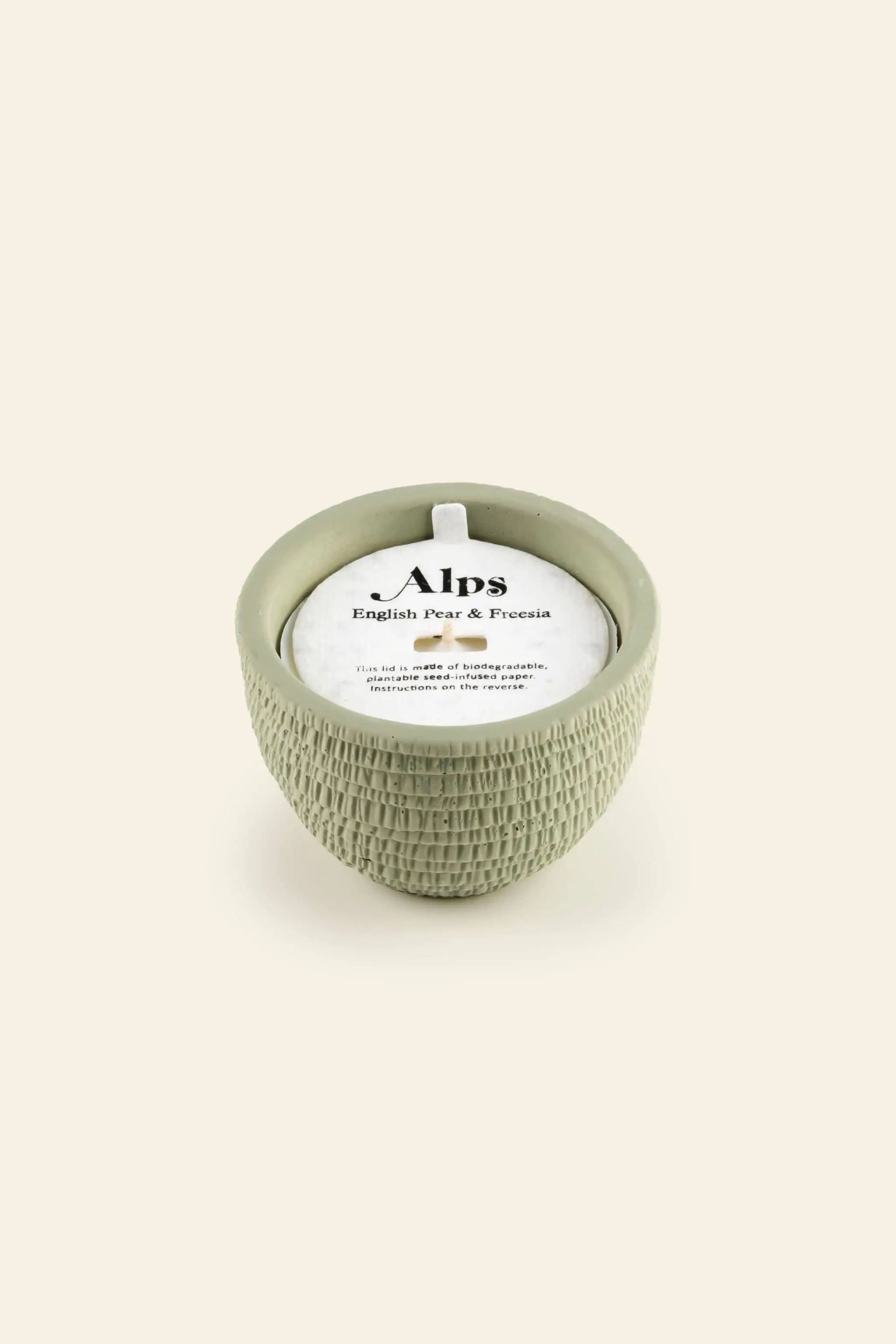 The Pass It On Alps Candle by Pass It On is a round, textured light-green holder with a white candle, featuring fresh pear and freesia scents on a plain cream background.