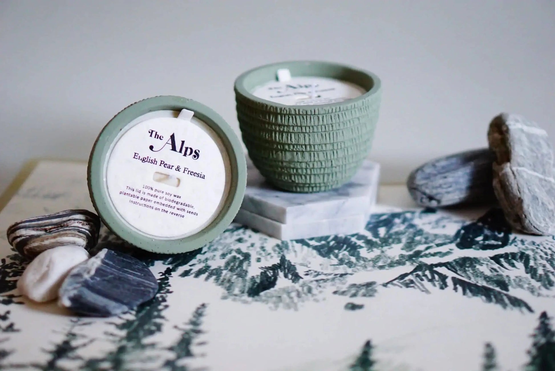A Pass It On Alps Candle from Pass It On, textured green with lid and labeled English Pear & Freesia, rests on a marbled coaster amid rocks and pine mountain art, filling the scene with notes of English Pear and Freesia.