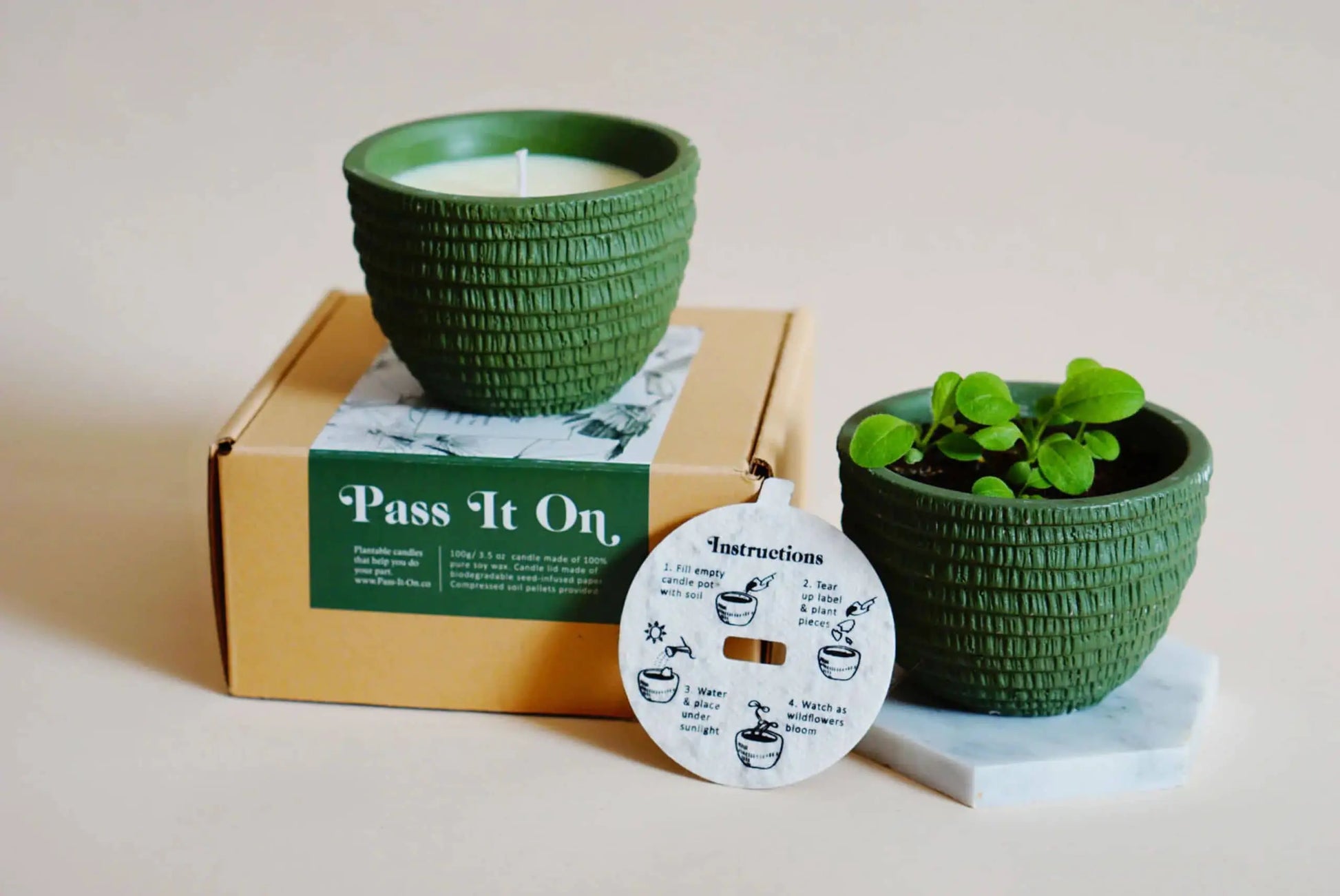 Two green textured pots are displayed: one is a Pass It On Alps Candle on a branded box, and the other holds a plant on a marble hexagon. An instruction card sits in front.