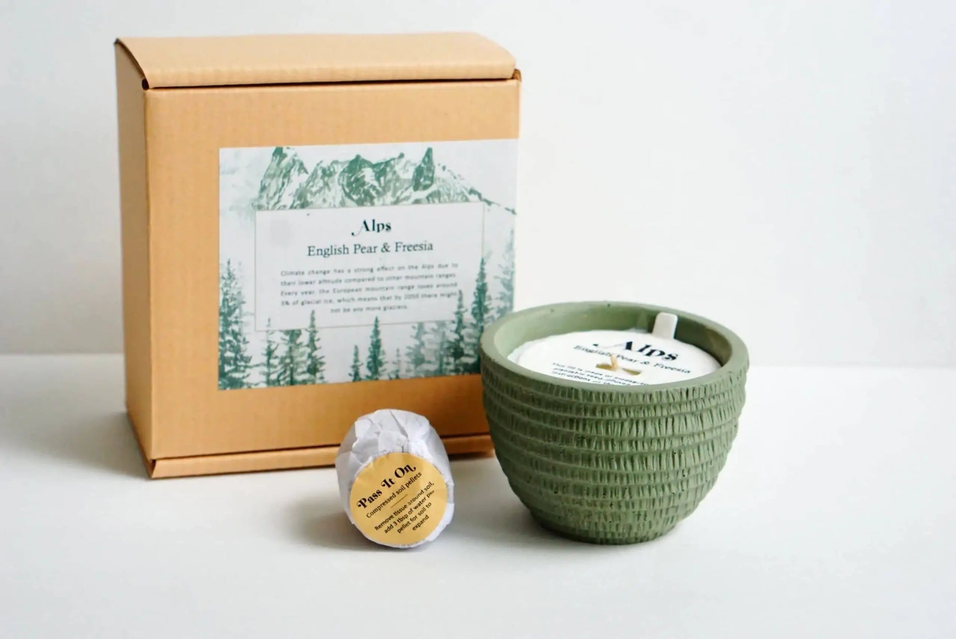 The Pass It On Alps Candle—a green textured candle in a ceramic bowl—sits next to its branded brown box and a “Peony & Citrus” wax melt with English Pear and Patchouli notes, all from Pass It On, arranged on a white background.