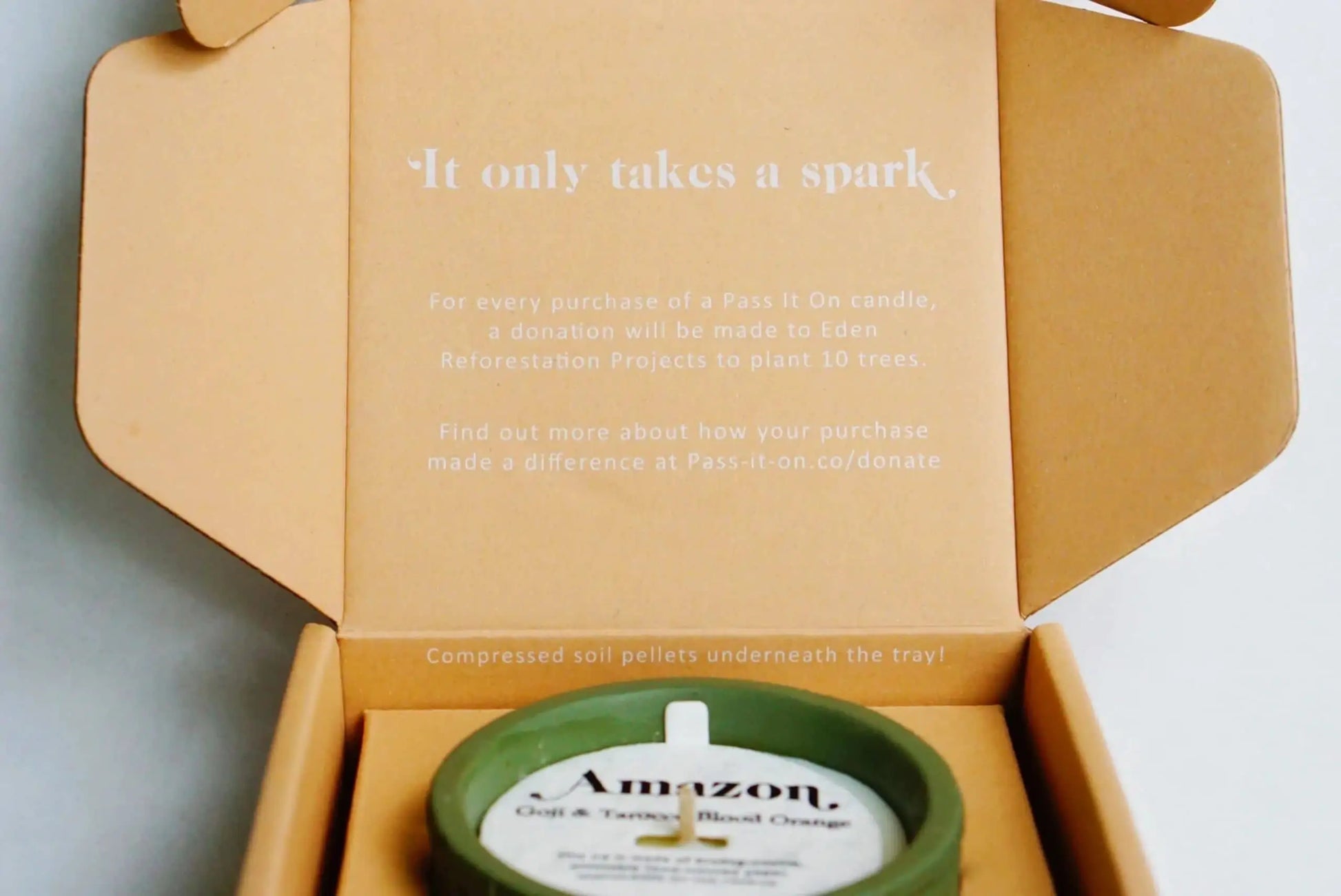 An open cardboard box reads: It only takes a spark. For every Pass It On Alps Candle (Freesia & Patchouli) purchase, Pass It On donates to Eden Reforestation Projects to plant 10 trees.
