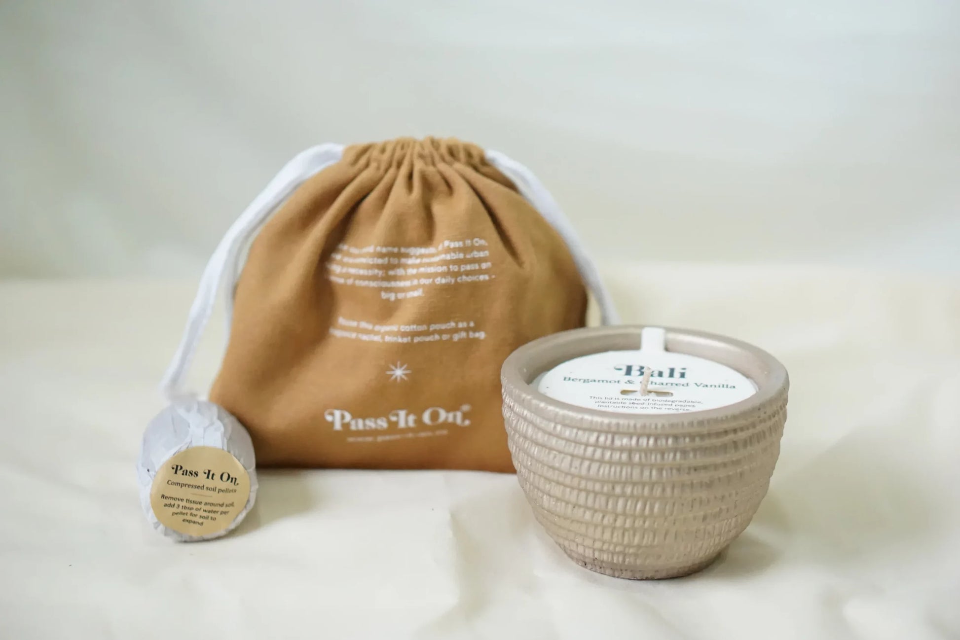 The Pass It On Bali Candle, labeled Bali, Bergamot & Burnt Vanilla, sits on a light surface with its brown drawstring Pass It On bag and a round tag, creating a soothing display from the Pass It On brand.