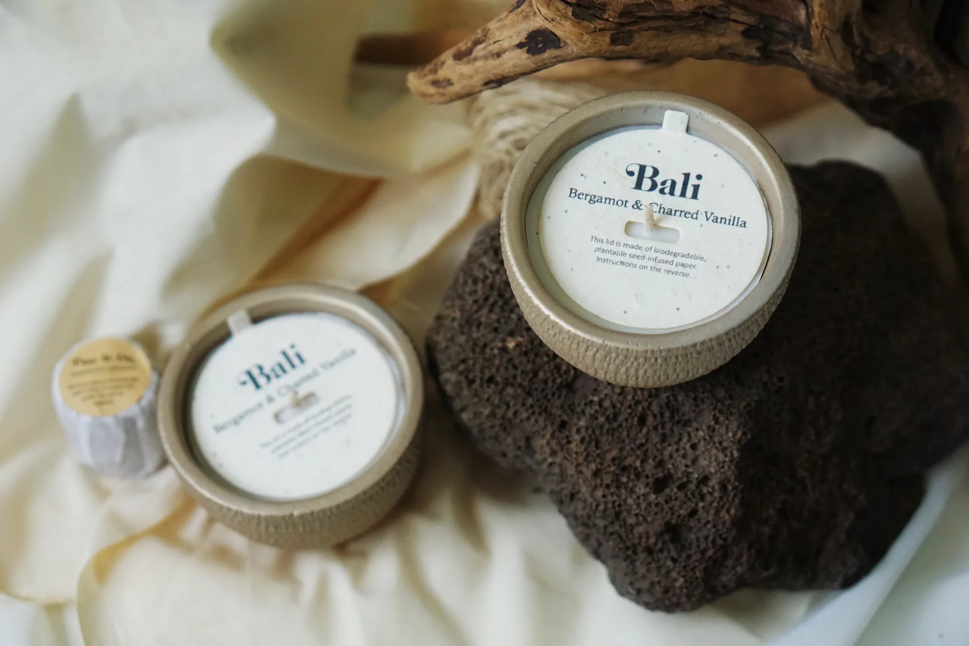 Three Pass It On Bali Candles rest on a textured rock with a wooden branch nearby, beautifully capturing the essence of Bali against a white cloth background.