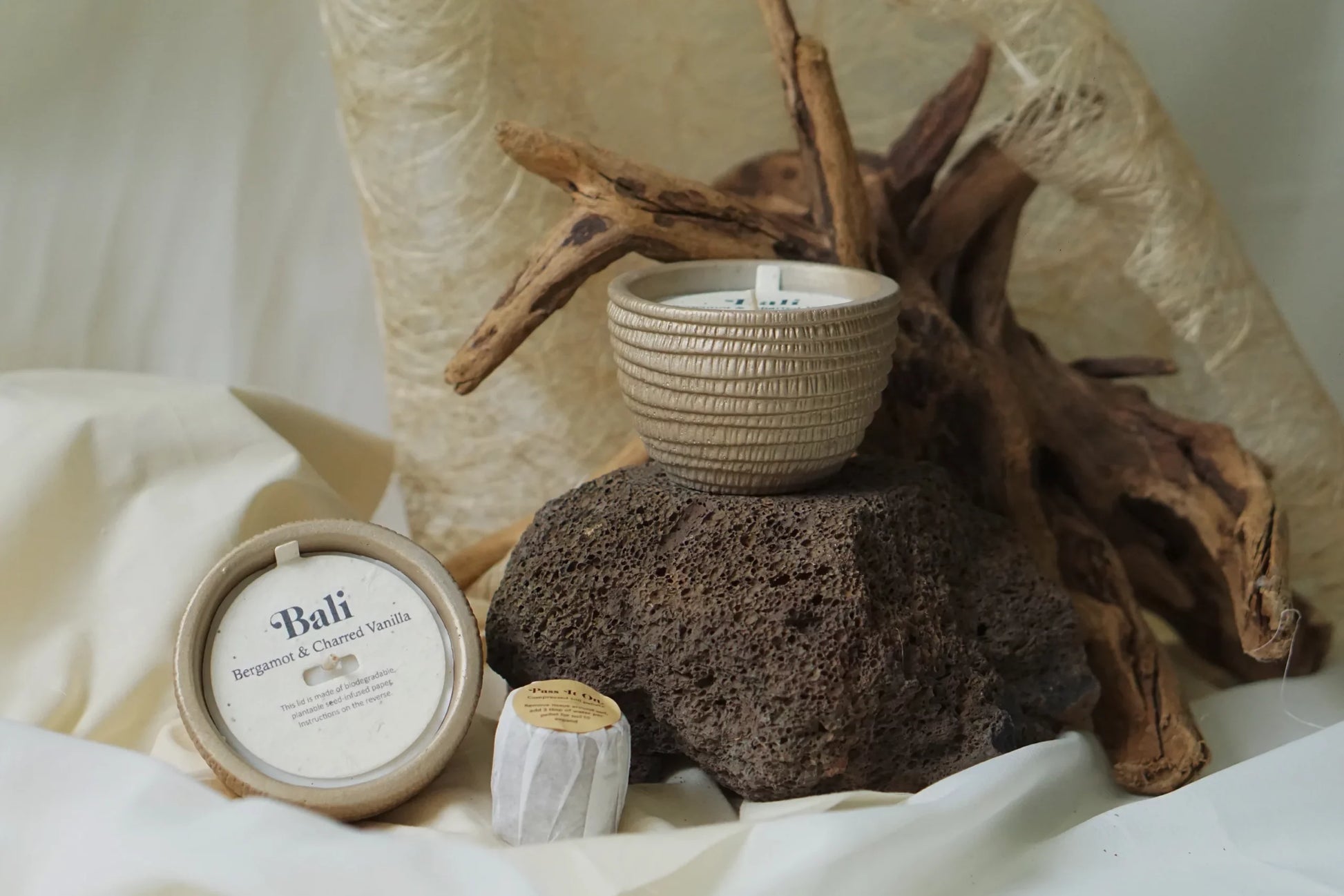 The Pass It On Bali Candle sits on a large volcanic rock with its labeled tin and a small wrapped item beside it. Driftwood and textured fabric create an earthy background, evoking the candle’s soothing vanilla and bergamot scent.