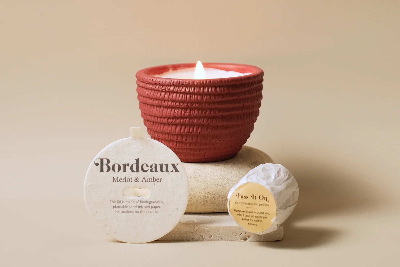 A Pass It On Bordeaux Candle in a textured red holder sits on stone with two round paper labels: one says “Bordeaux Merlot & Amber,” capturing rich wine notes, and the other reads “Pass it On.” The scene features a soft beige background.