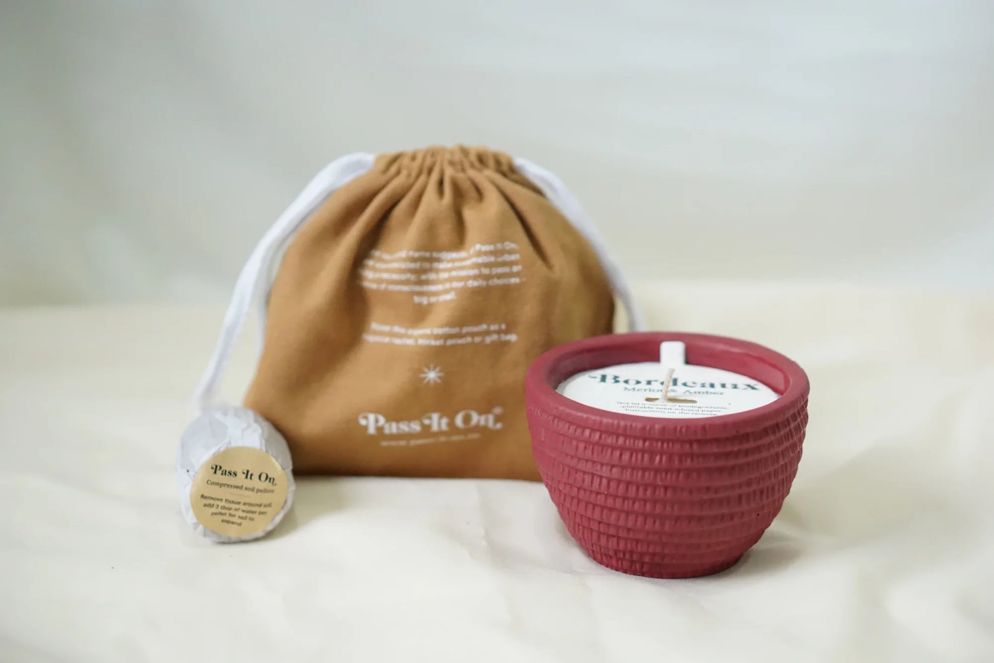 A small brown drawstring bag, a wrapped Pass It On item, and a red textured Pass It On Bordeaux Candle are displayed on a light fabric surface.