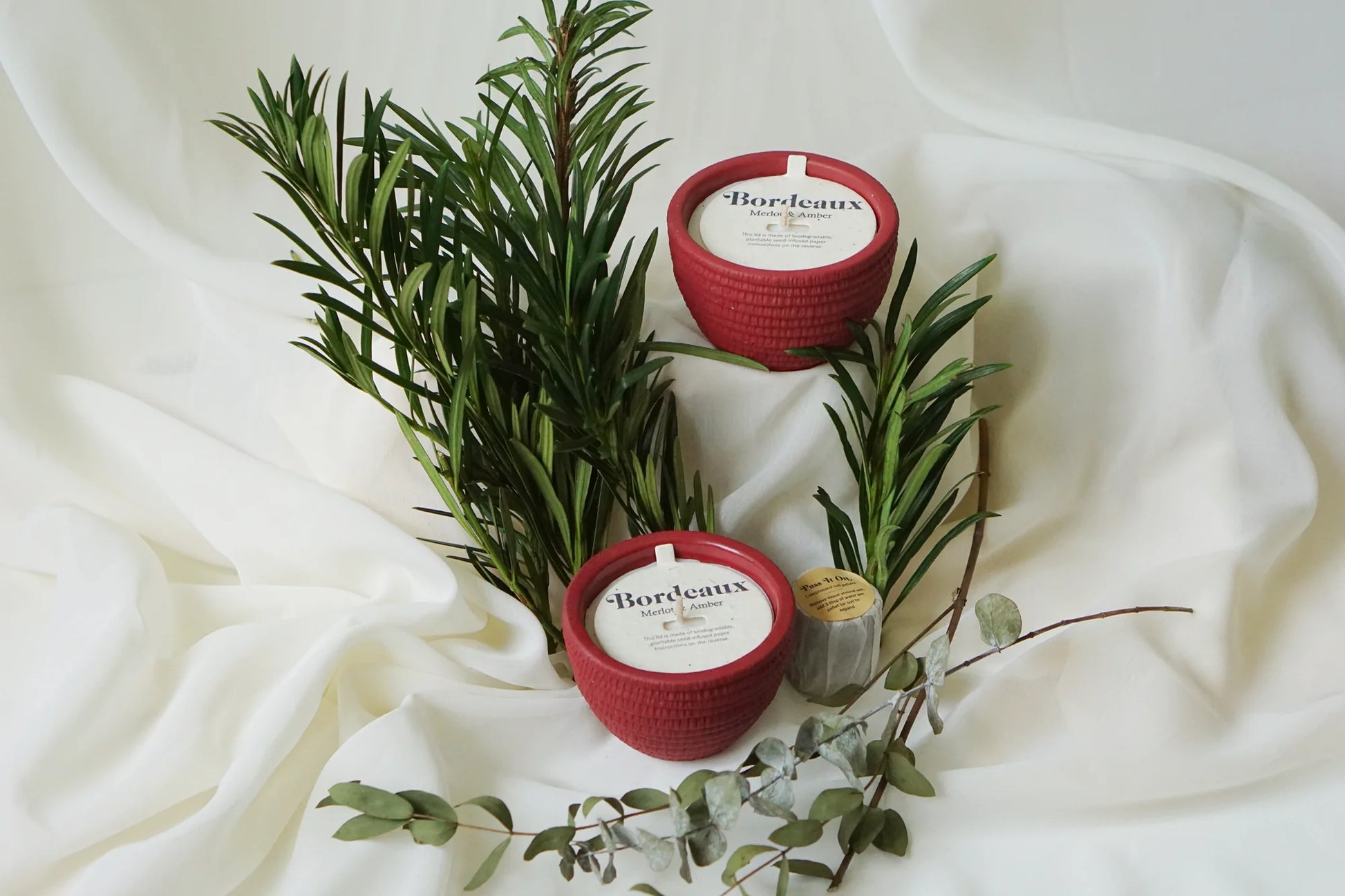 Two Pass It On Bordeaux Candles rest on white fabric, surrounded by green foliage and eucalyptus, for a fresh and elegant look that evokes the rich aroma of Merlot.
