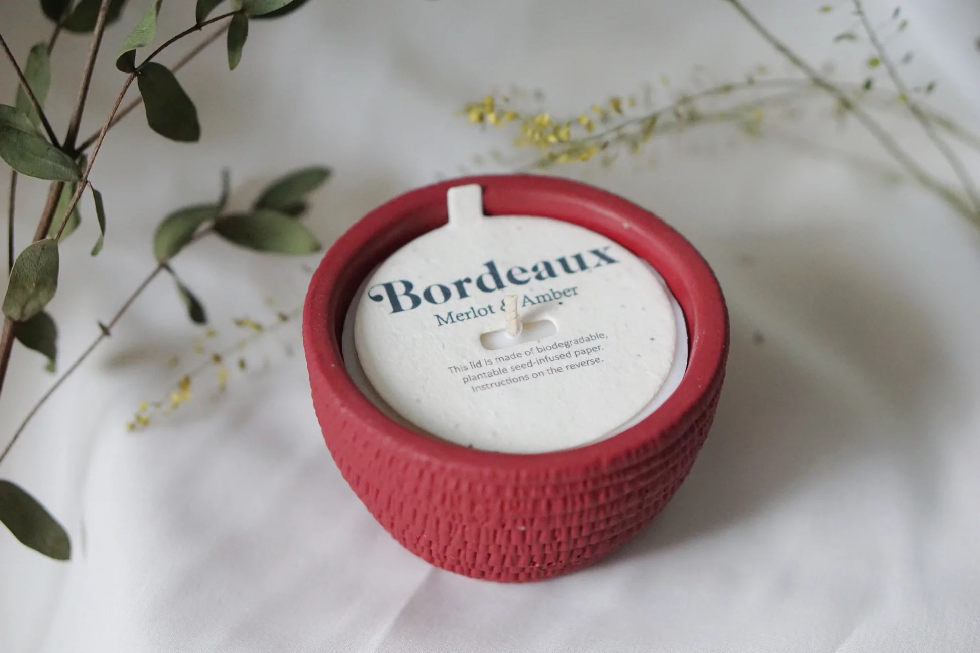 A round red Pass It On Bordeaux Candle with a textured exterior sits on a white surface amid greenery. The candle, labeled with white, blue, and gold text, evokes rich red wine and black cherry scents. Brand: Pass It On.