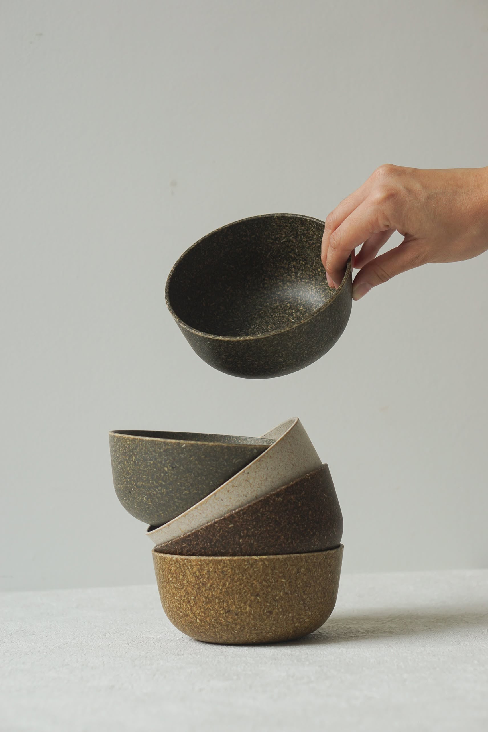 A hand lifts the Pass It On Capsule Bowl, Rice Husk—an earthy-toned bowl crafted from upcycled rice husks—from a stack of four similar bowls by Pass It On, set against a plain light background.