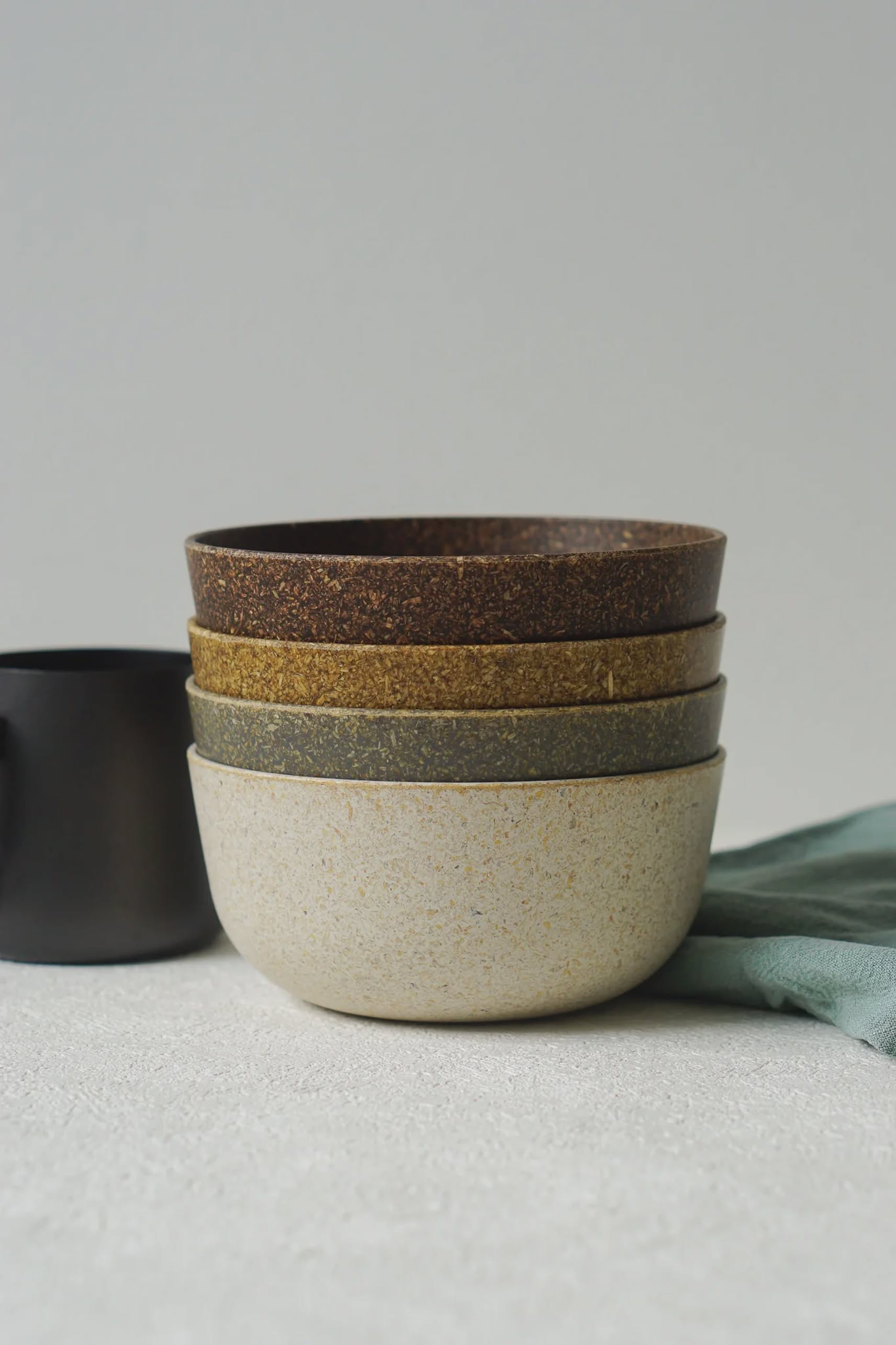 A stack of four Pass It On Capsule Bowls, made from upcycled rice husks in neutral tones, are arranged largest to smallest beside a black mug and blue cloth on a textured white surface.