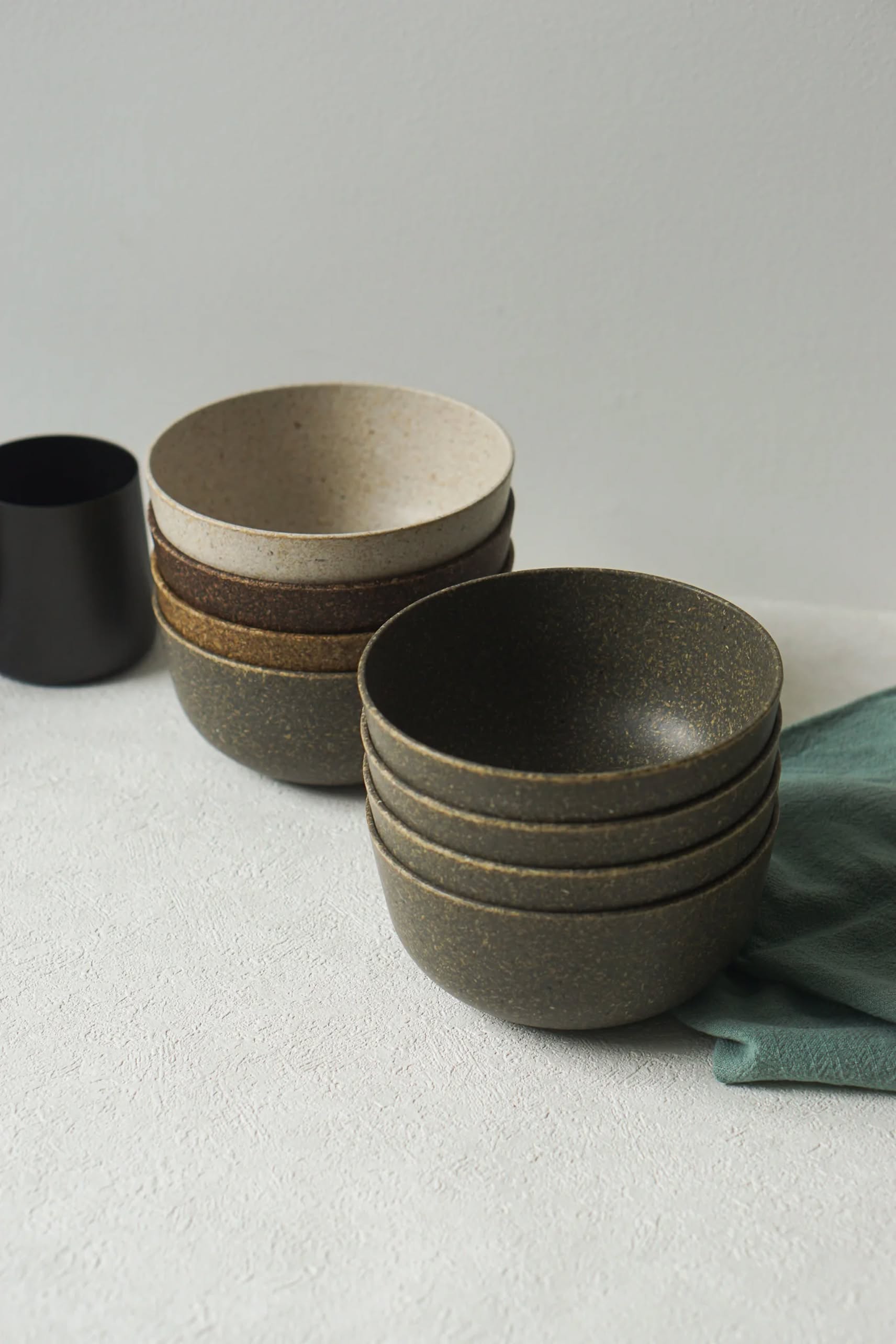 Two stacks of Pass It On Capsule Bowl, Rice Husk ceramic bowls, made from upcycled rice husks in earth tones, rest on a textured white surface with a green cloth and black cup against a light gray background, showcasing Pass It On’s eco-conscious design.