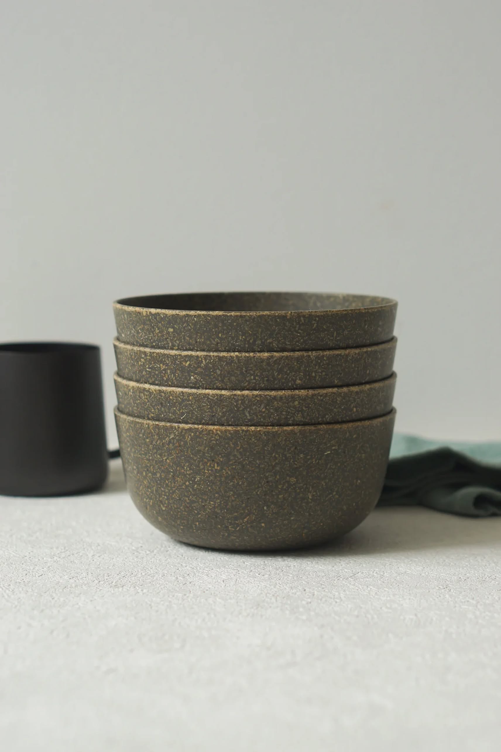 Four stacked, speckled brown Pass It On Capsule Bowls made from upcycled rice husks rest on a light surface, with a dark cup and green cloth in the background—showcasing sustainable style by Pass It On.