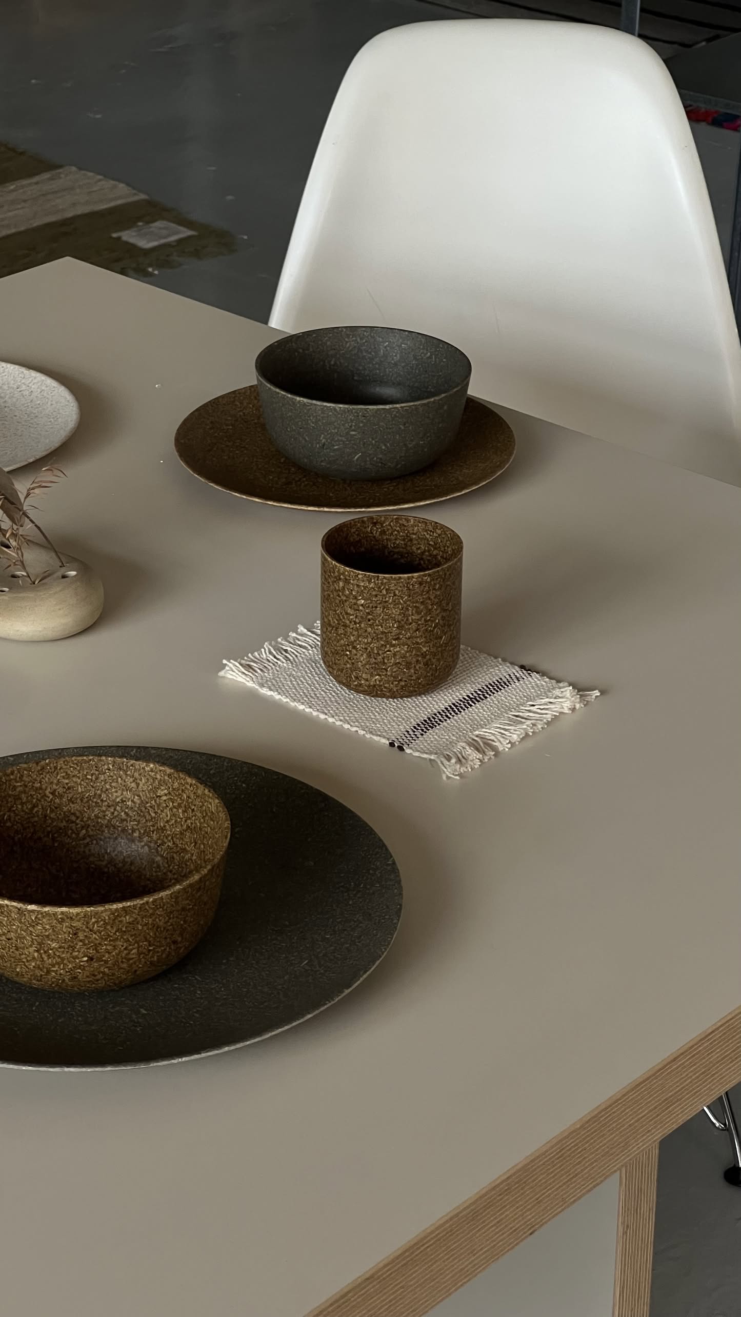 A minimalist table with dark ceramic plates, bowls, a Pass It On Capsule Cup (Rice Husk) on a small woven mat, and a decorative vase on a light table beside a white chair for an eco-friendly touch.