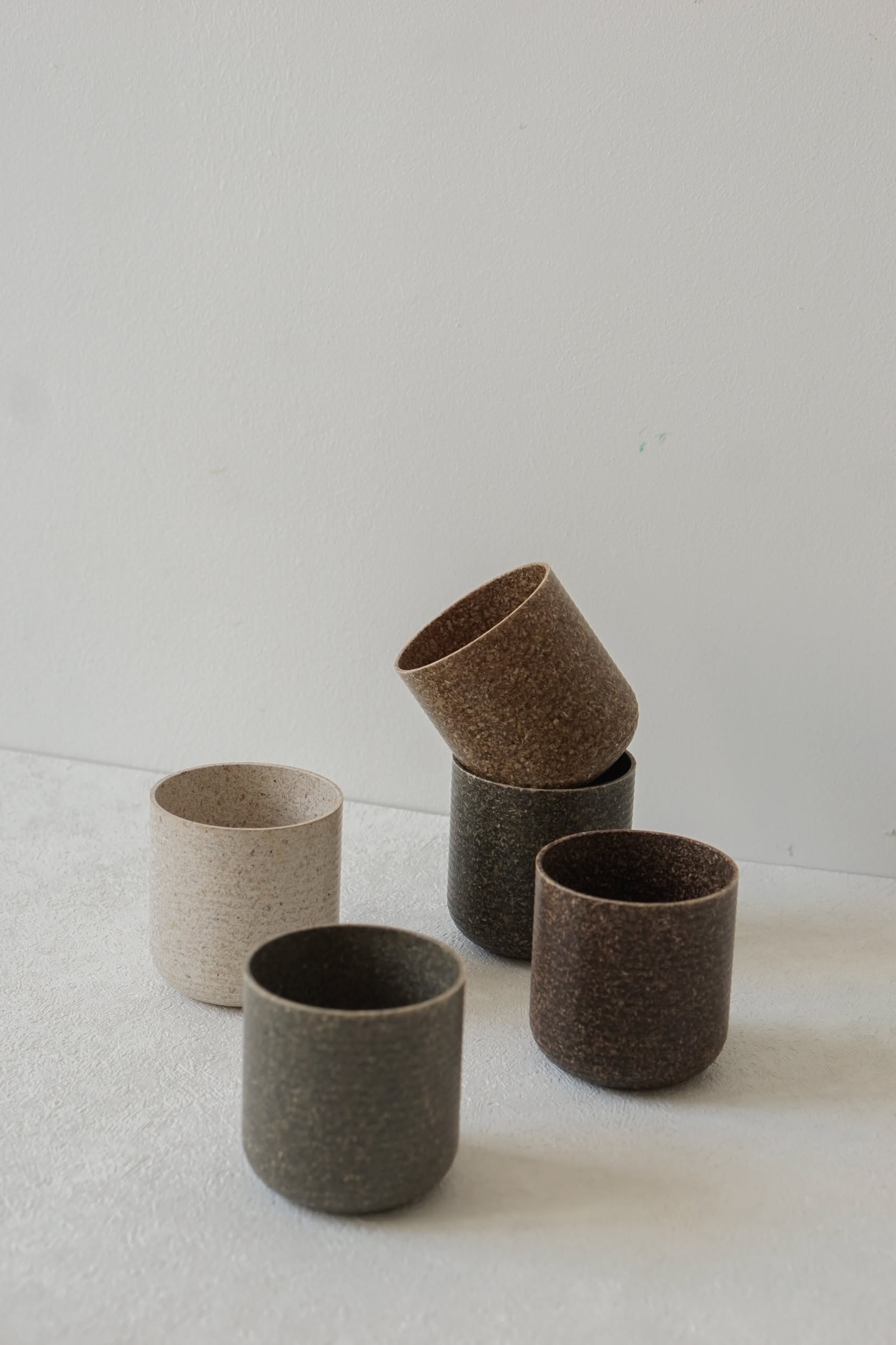 Five Pass It On Capsule Cups, made from upcycled rice husks, are arranged on a light surfaceâtwo stacked and three separateâin earthy tones. These eco-friendly cups by Pass It On sit against a plain, neutral background.
