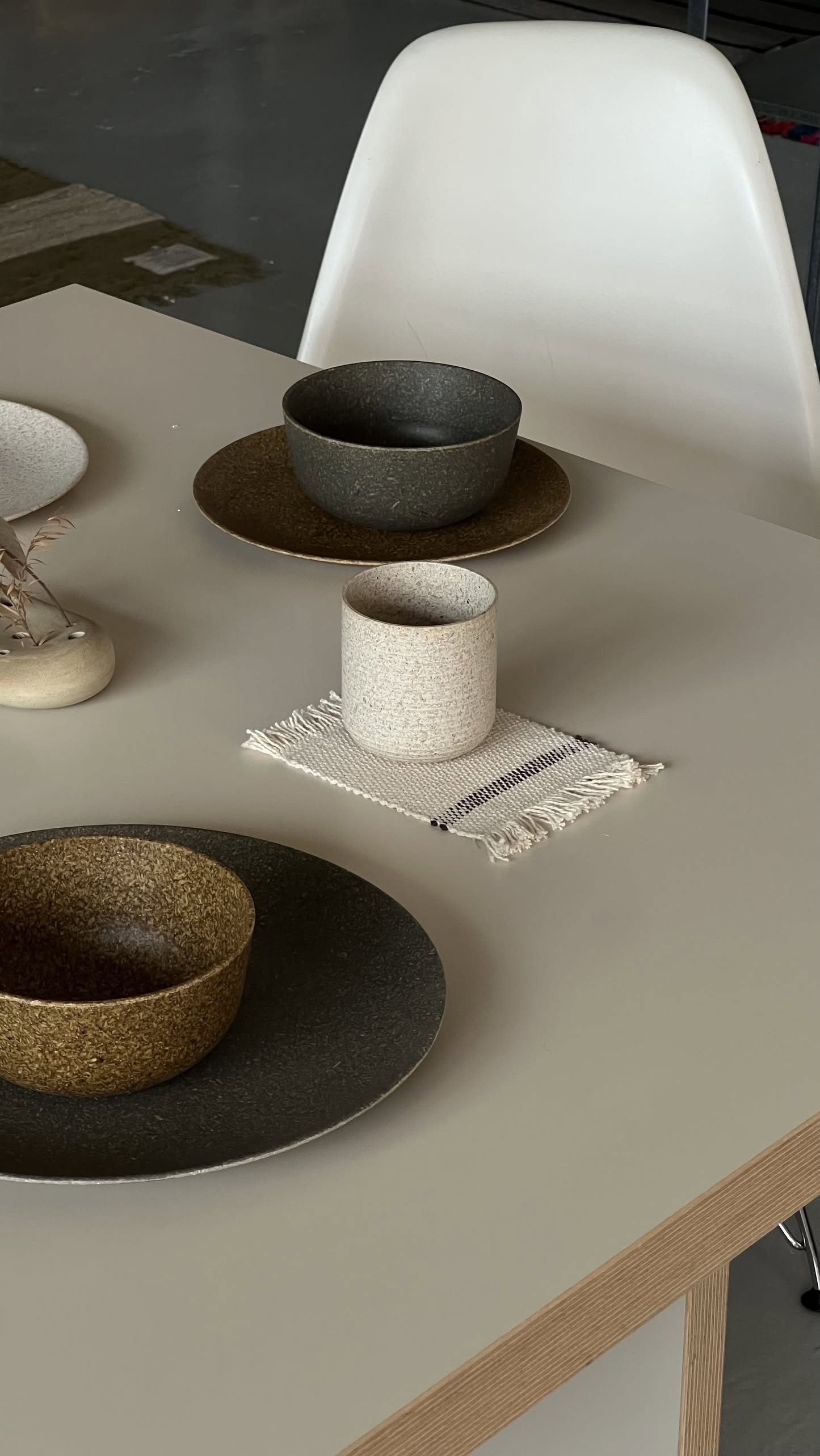 A minimalist dining table set features textured ceramic bowls and plates in earth tones, a small beige vase with dried grass, and the Pass It On Capsule Cup made from upcycled rice husks by Pass It On on a woven coaster. A white chair is at the table.