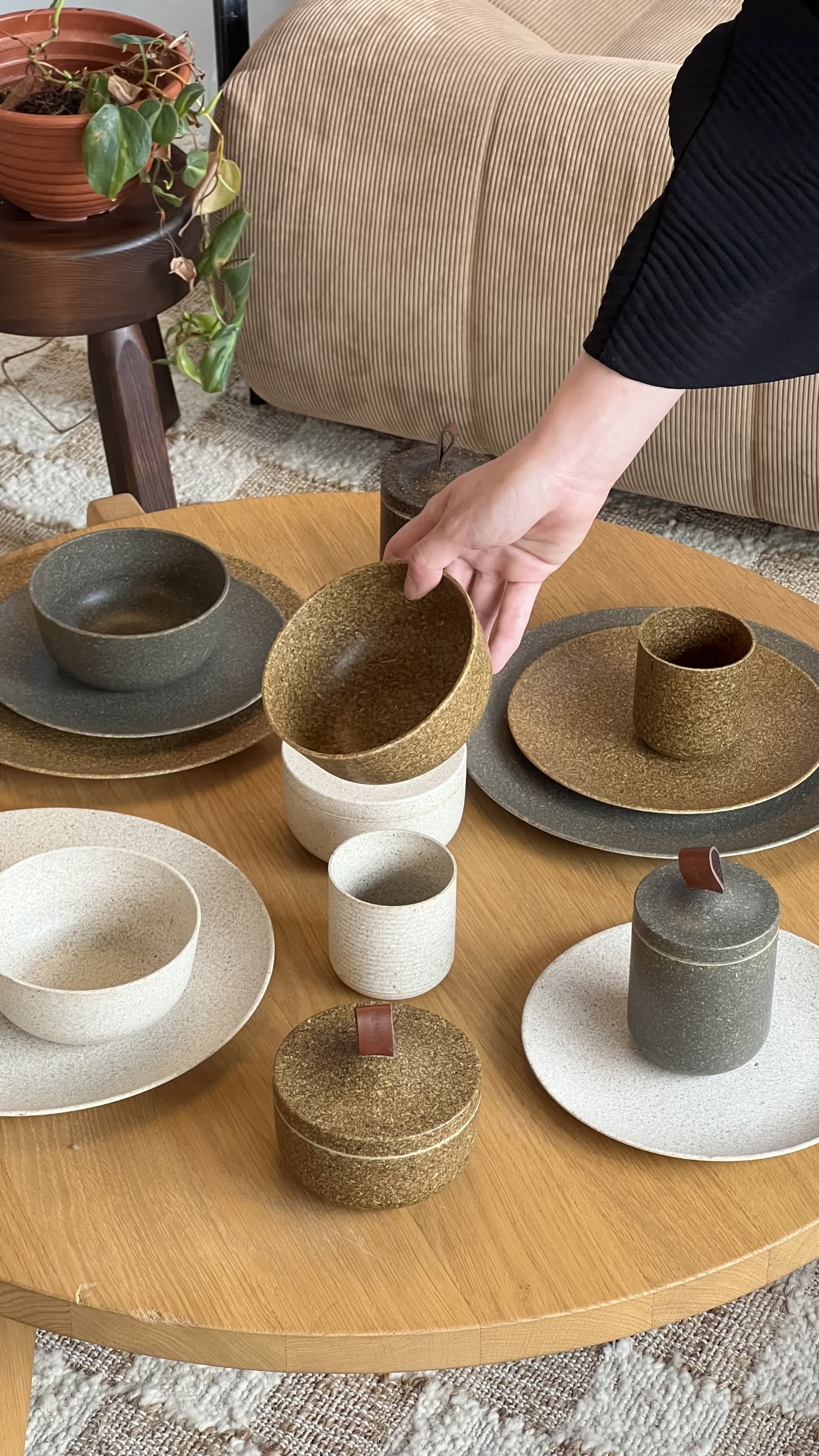 A hand sets a speckled bowl on a wooden table arranged with ceramic dishes, cups, and bowlsâincluding the Pass It On Capsule Cup made from upcycled rice husks. A plant and textured furniture appear in the background.