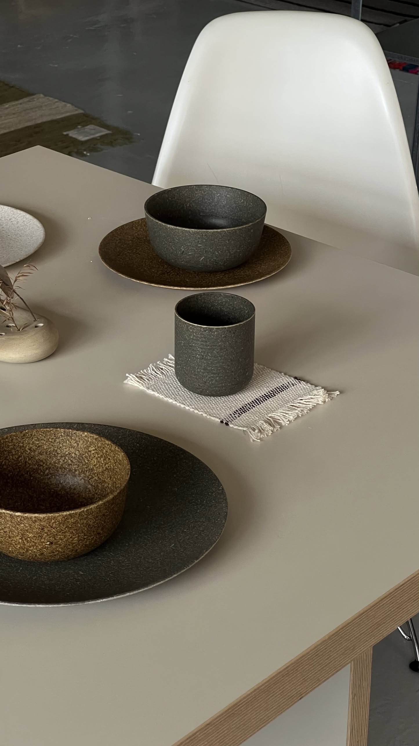 A modern dining table set with minimalist dishware, featuring two textured bowls on matching plates and a Pass It On Capsule Cup, Rice Husk by Pass It On on a small woven mat, next to a white chair. Neutral and earthy tones dominate the scene.