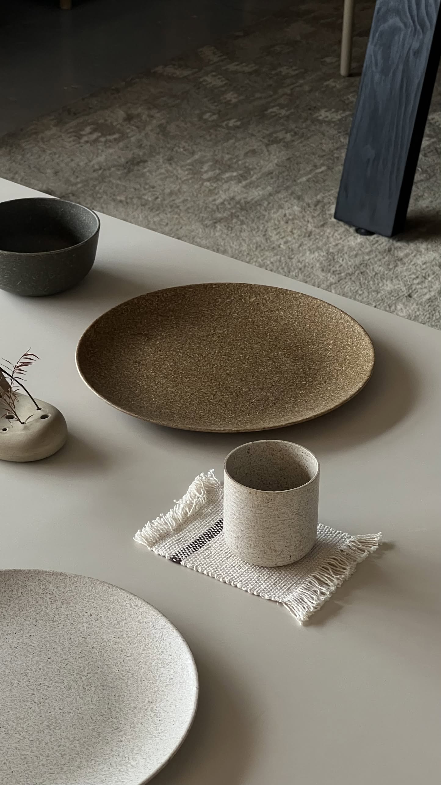 A minimalist table setting featuring Pass It On’s Dining Plate L in Rice Husk, a beige cup on a fringed cloth, a small gray bowl, and a ceramic vase with dried flowers—all arranged on a light surface for an eco-conscious look.